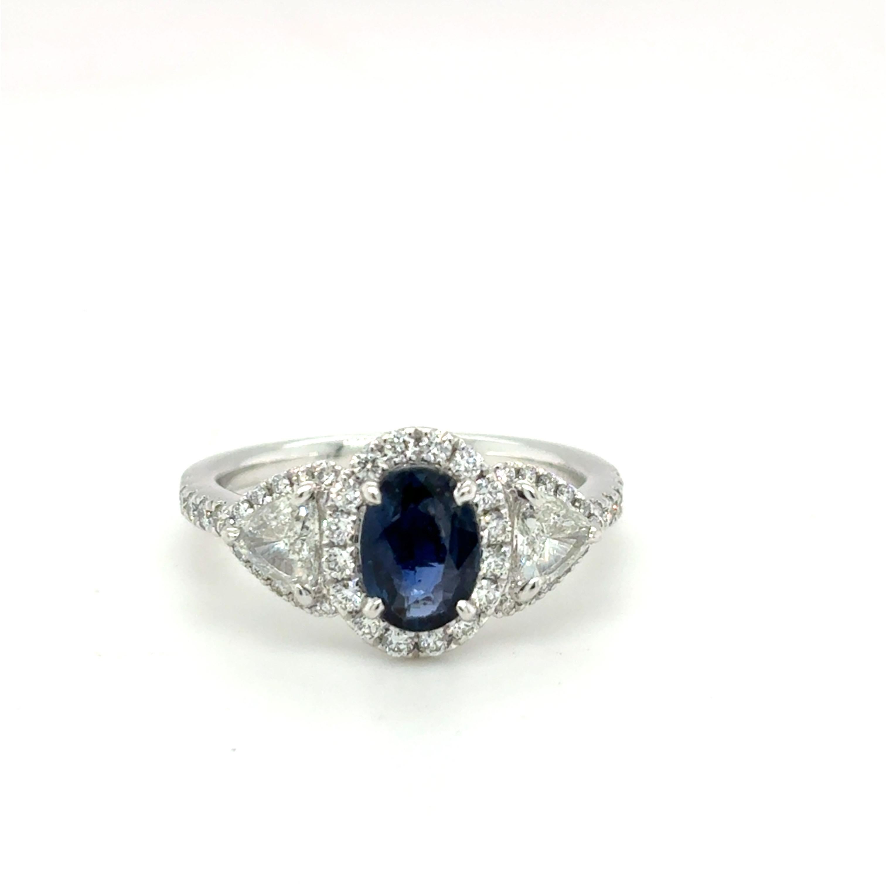 1.183carat oval cut blue sapphire ring, framed by a sparkling halo of white diamonds and flanked by two 0.53 carat trillion cut diamonds on the east and west sides. Expertly crafted in platinum using VS1 clarity and G-H color diamonds. Ring size is