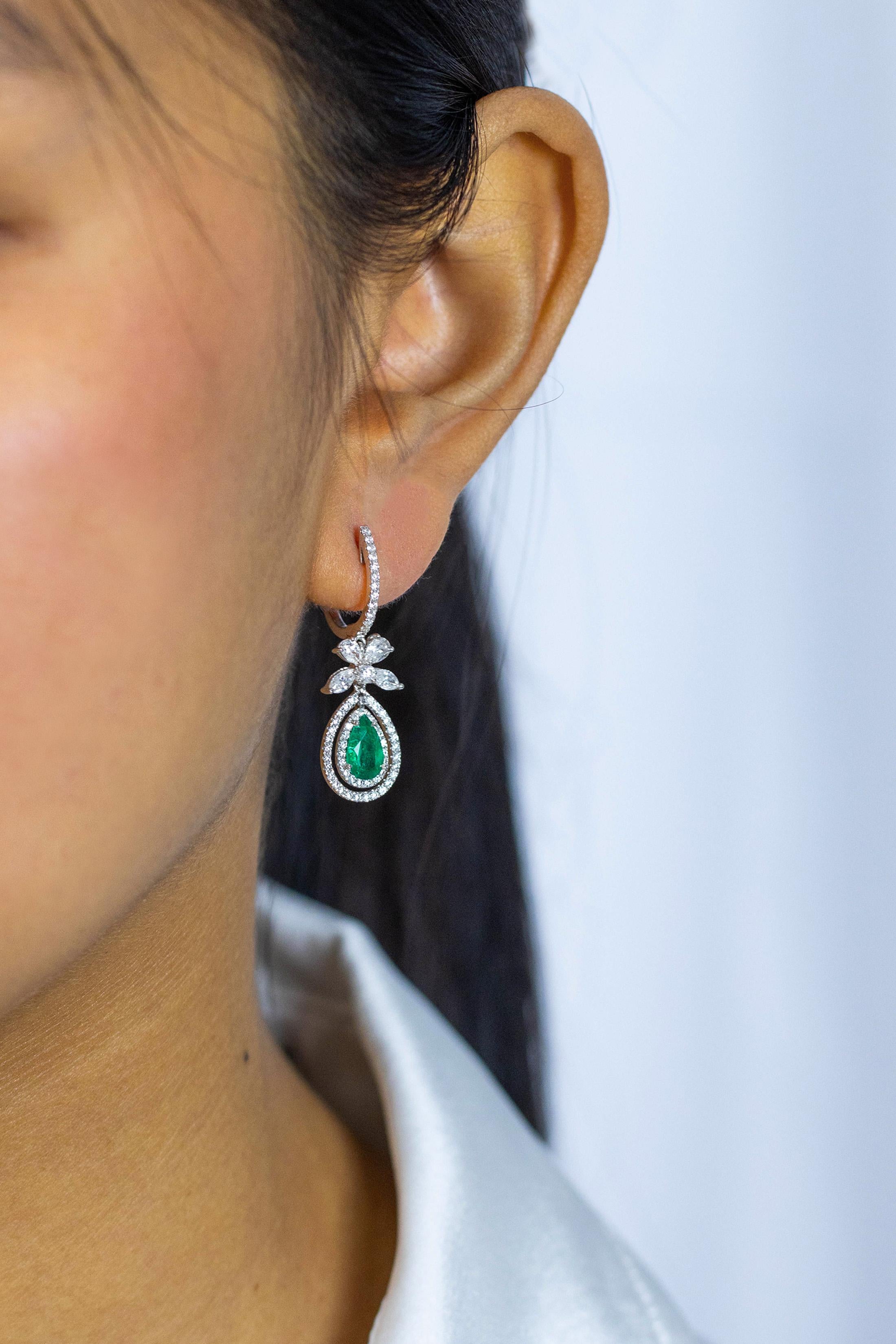 These gorgeous earrings feature a pear shaped green emerald center stone weighing 1.14 carats with 2 rows of brilliant round diamonds that go around the emerald. A bow-like figure made with diamonds hang on top of the emeralds and even more round