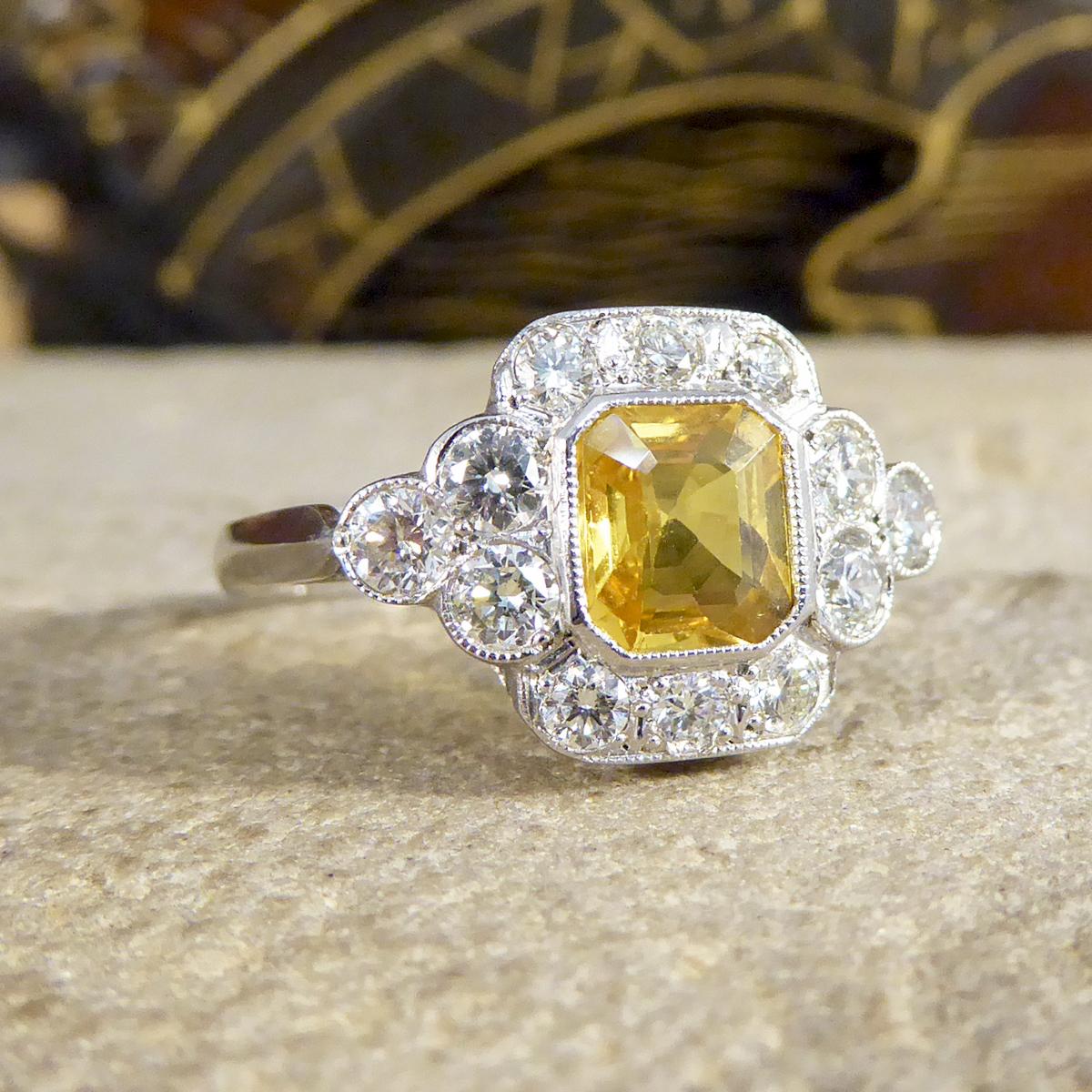 This fabulous Contemporary cluster ring dazzles on the hand and has been crafted to resemble an Edwardian bordering Art Deco style. It features a single central Asscher Cut Yellow Sapphire stone weighing 1.15ct, surrounded by 12 Brilliant cut