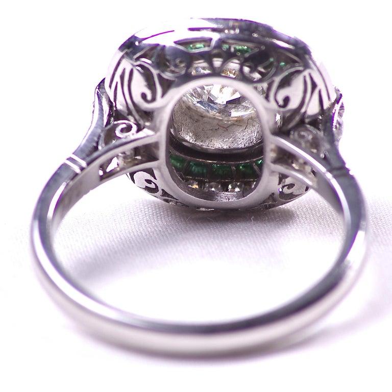 An intricate double halo with vibrant colors creates this vintage inspired engagement ring. Featuring a 1.18 carat old mine cut diamond that is J color, SI2 clarity surrounded by celestial circles of glowing forest green emeralds and numerous old