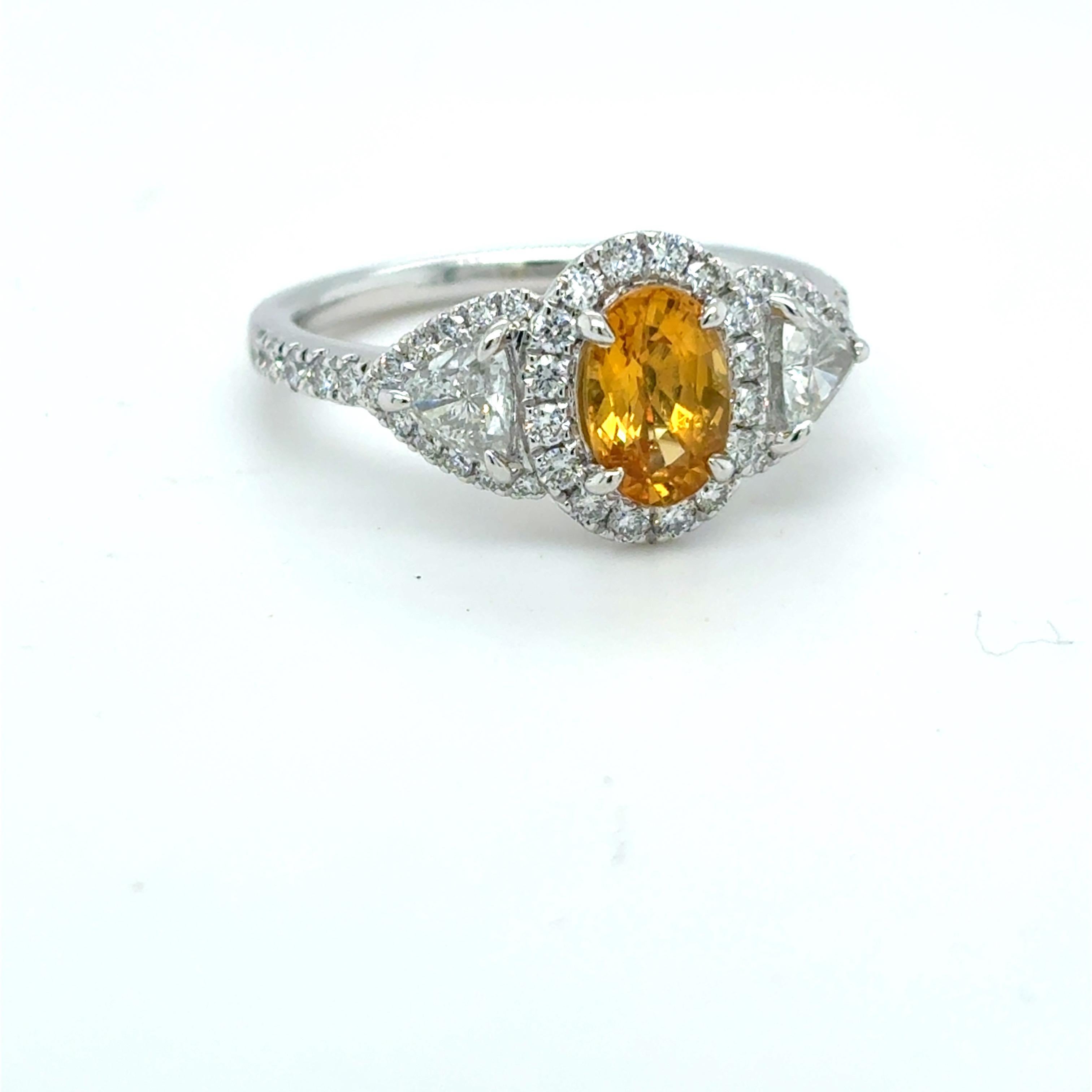A captivating 1.18 carat oval cut yellow sapphire takes center stage in this exquisite platinum ring, encircled by a dazzling halo of fine white diamonds. Flanked by two perfectly matched 0.50 carat trillion cut diamonds on either side, the design