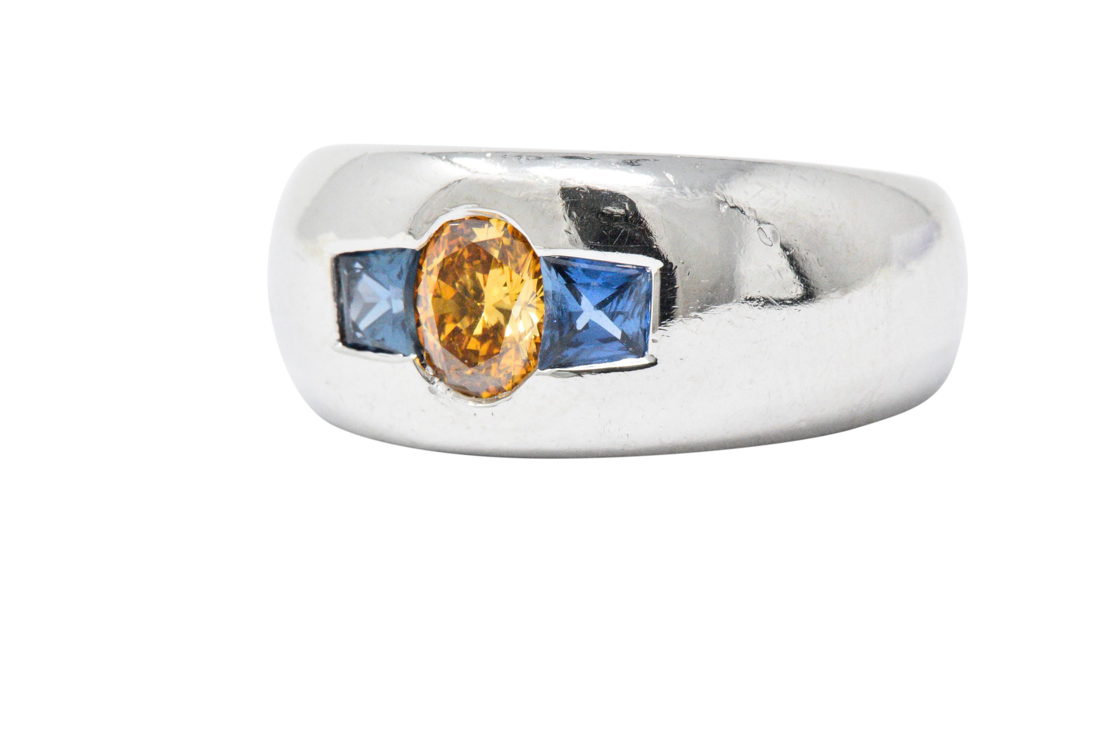 Centering an oval cut diamond weighing 0.59 carats, natural fancy intense yellow-orange color and VS1 clarity

Flanked by two trapezoid cut sapphires weighing approximately 0.60 carats total, bright, deep cornflower blue

In a flush or 