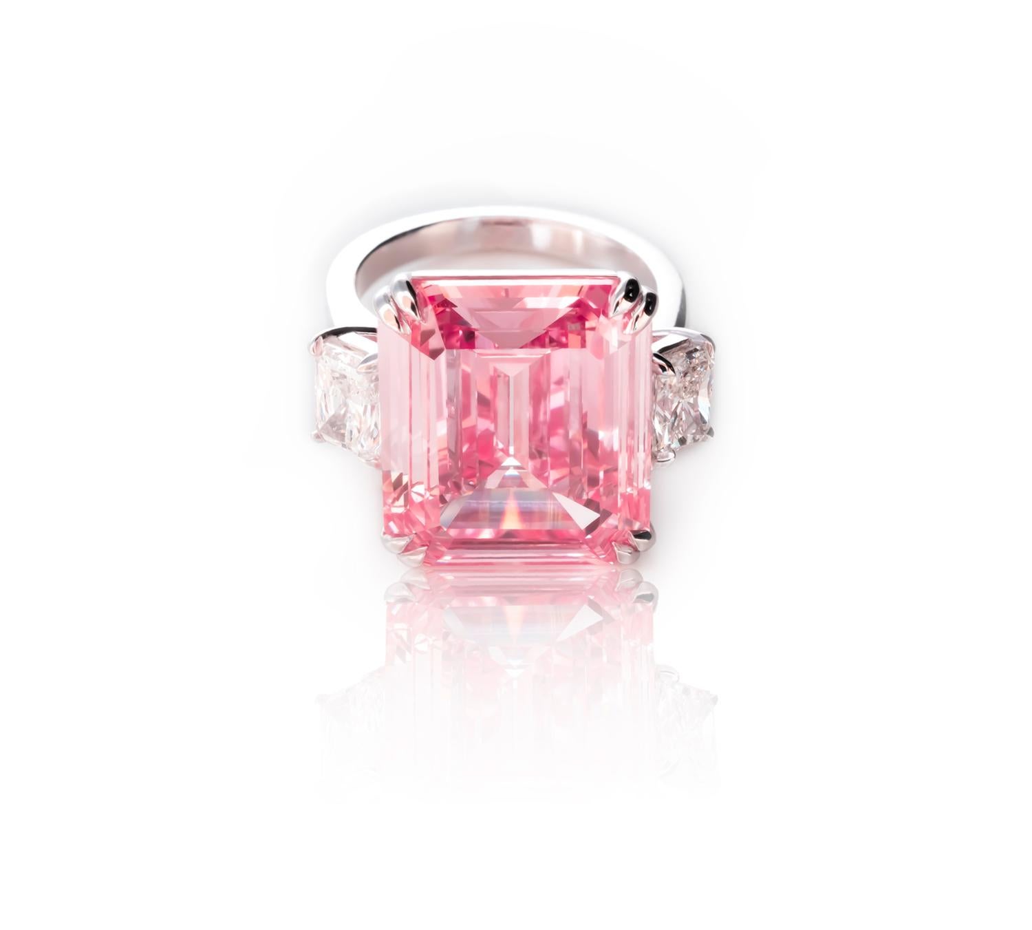 This unparalleled ring showcasing a stunning Emerald Cut 12-carat pink diamond with desirable bubble gum shades is an absolute rarity. The pink diamond is embraced by sparkling radiant-cut white diamonds on either side, creating a contrast that