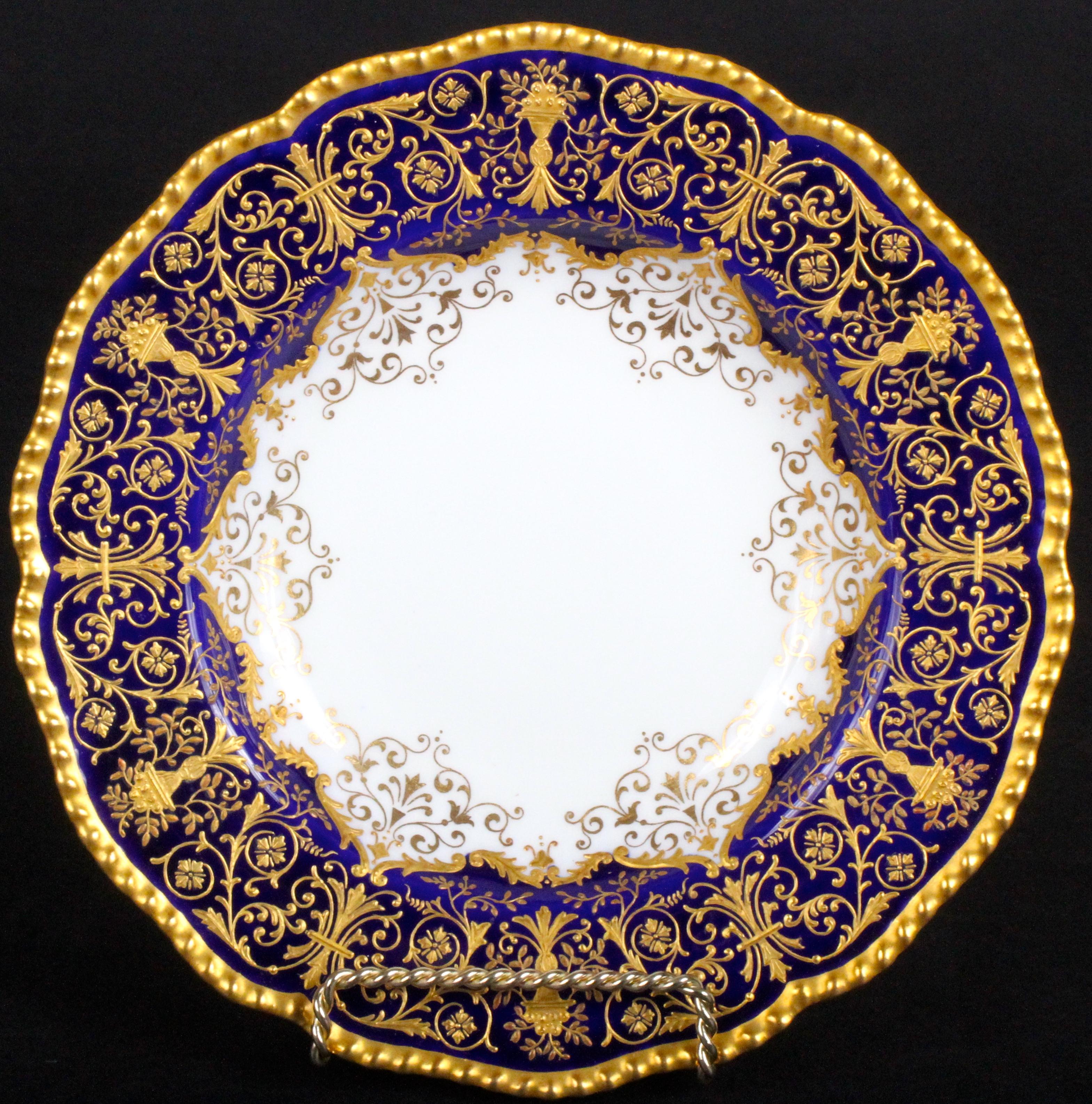 These 12 ornate soup plates are from the esteemed firm of Coalport, Shopshire, England. The soup plates feature wonderfully intricate raised-paste gold work of alternating ornaments of 2 different designs, one of a grand vase spewing meandering