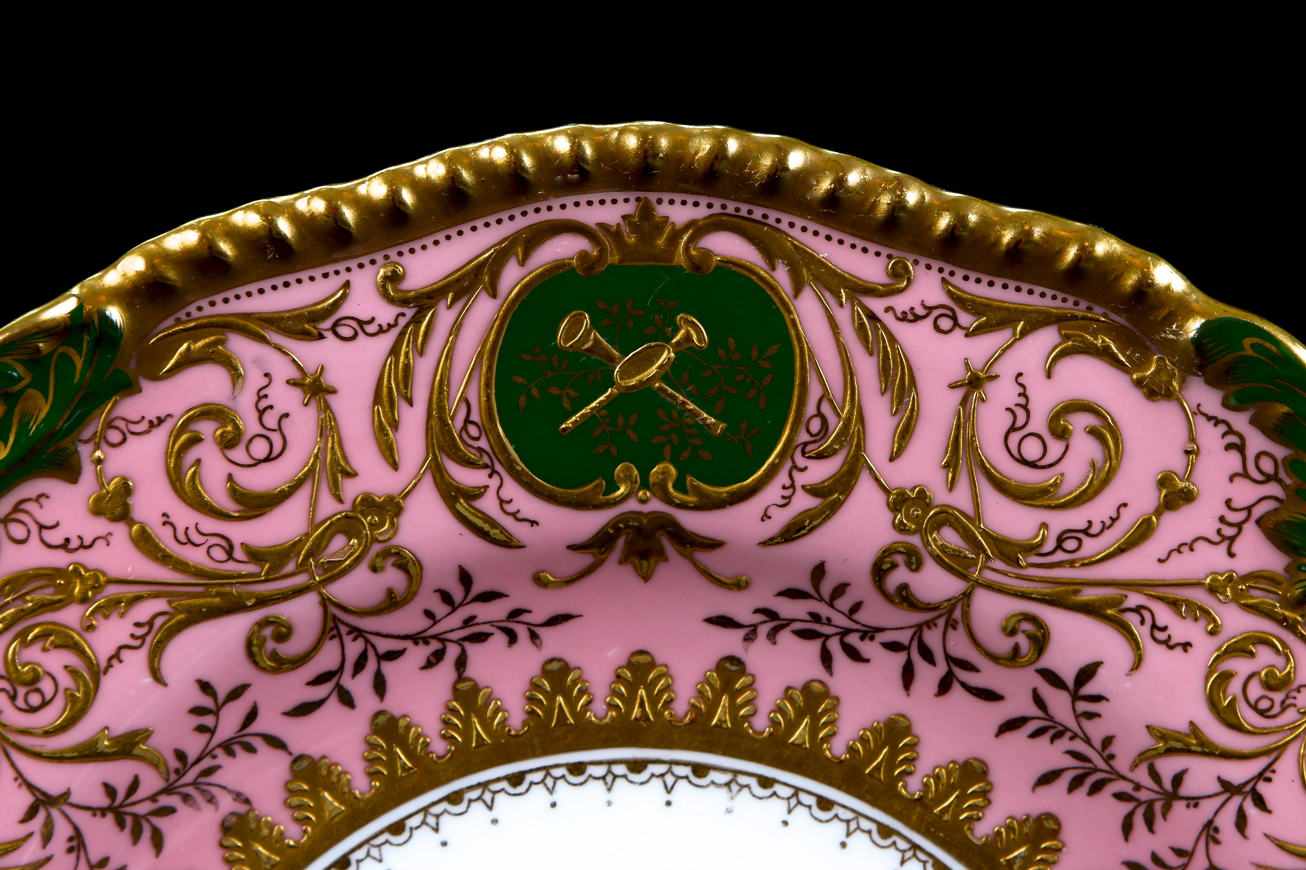 These 19th century plates from Coalport, England feature pink ground with an elaborate scrolling foliate gilded design, accented by 3 cartouches or inserts of musical instruments on a green background. The gilding on these plates is thick and