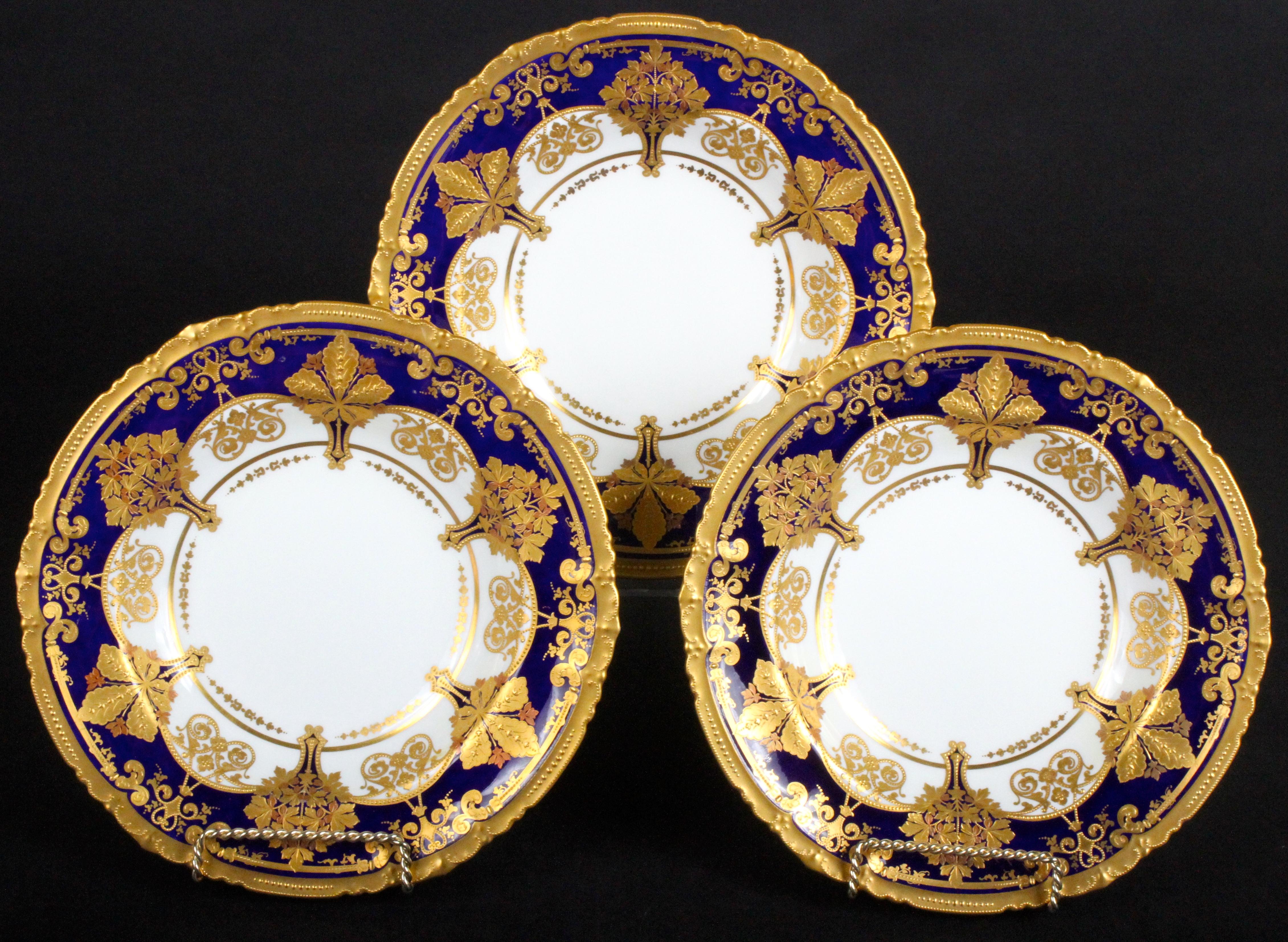 These 12 ornate soup plates are from the esteemed Royal Crown Derby firm of Derby, England. The soup plates' cobalt rim features 6 ornaments of 2 different designs, both composed of a variety of leaves in yellow and rose gold, the soup plates are
