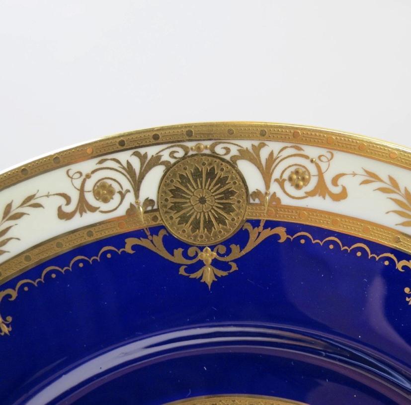 A classic and elegant set of 12 Minton England bone china dinner plates. This set features a nice deep collar of cobalt blue that is accented with raised tooled gilded swags and their signature gold medallions on their collars. Made of crisp white