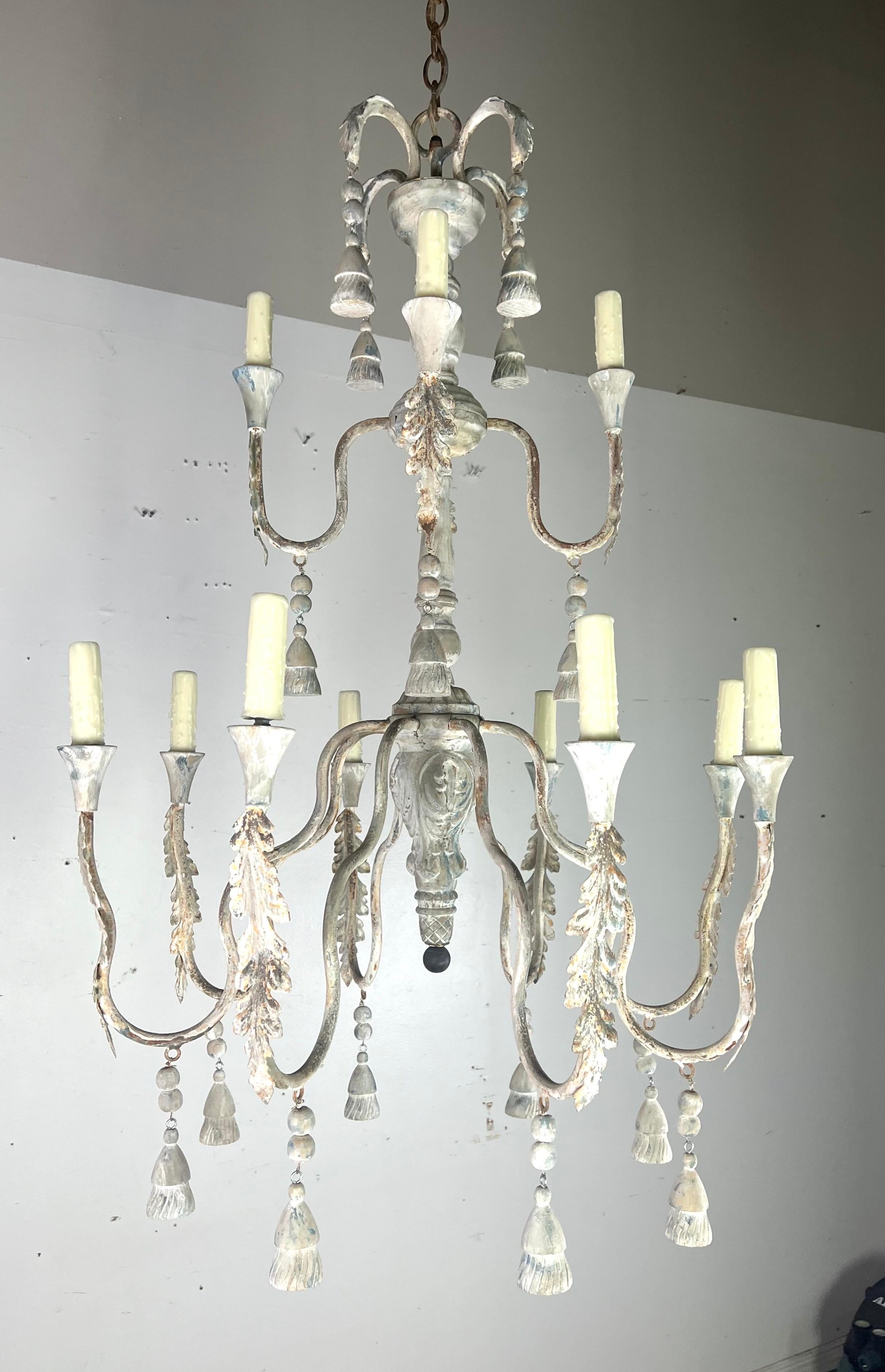 12-light Tuscan style two-tier painted chandelier by Melissa Levinson.  It features a multi-tiered design with twelve curved arms, each holding candle-like lights with drip wax candle covers.  The chandelier is adorned with acanthus leaves along the