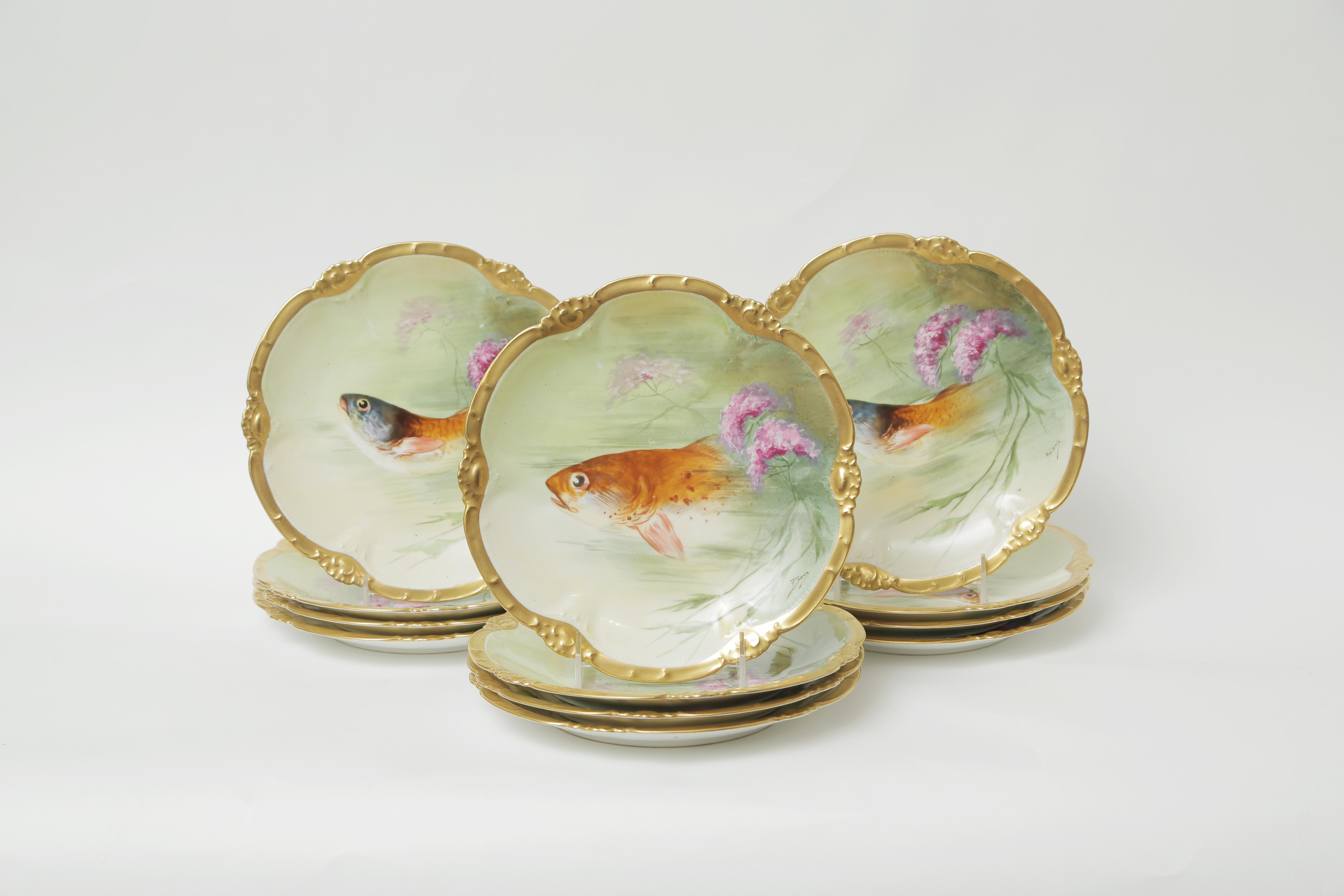 Un ensemble vibrant de 12 assiettes à poisson de Limoges France. Une assiette de forme agréable, rehaussée d'or 24 carats et entièrement peinte à la main avec des motifs de la flore et de la faune. Les couleurs et le design sont superbes et l'objet