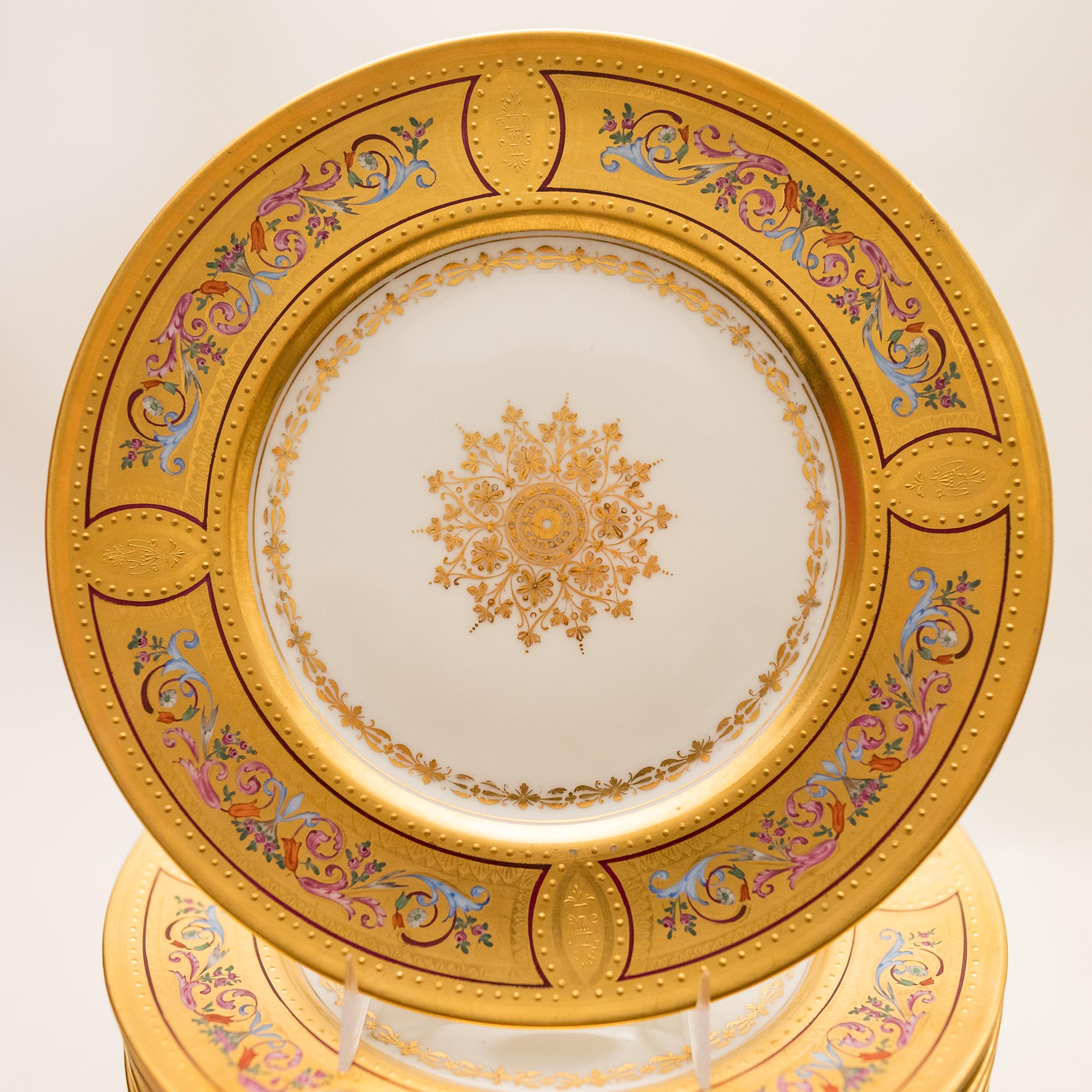 An exquisite set of dessert plates featuring colorful and detailed enamel work on their gilded collars. Beautiful swags of florals and urns in vibrant enameling with double ruby banding. They also feature a gilded snowflake center medallion. In