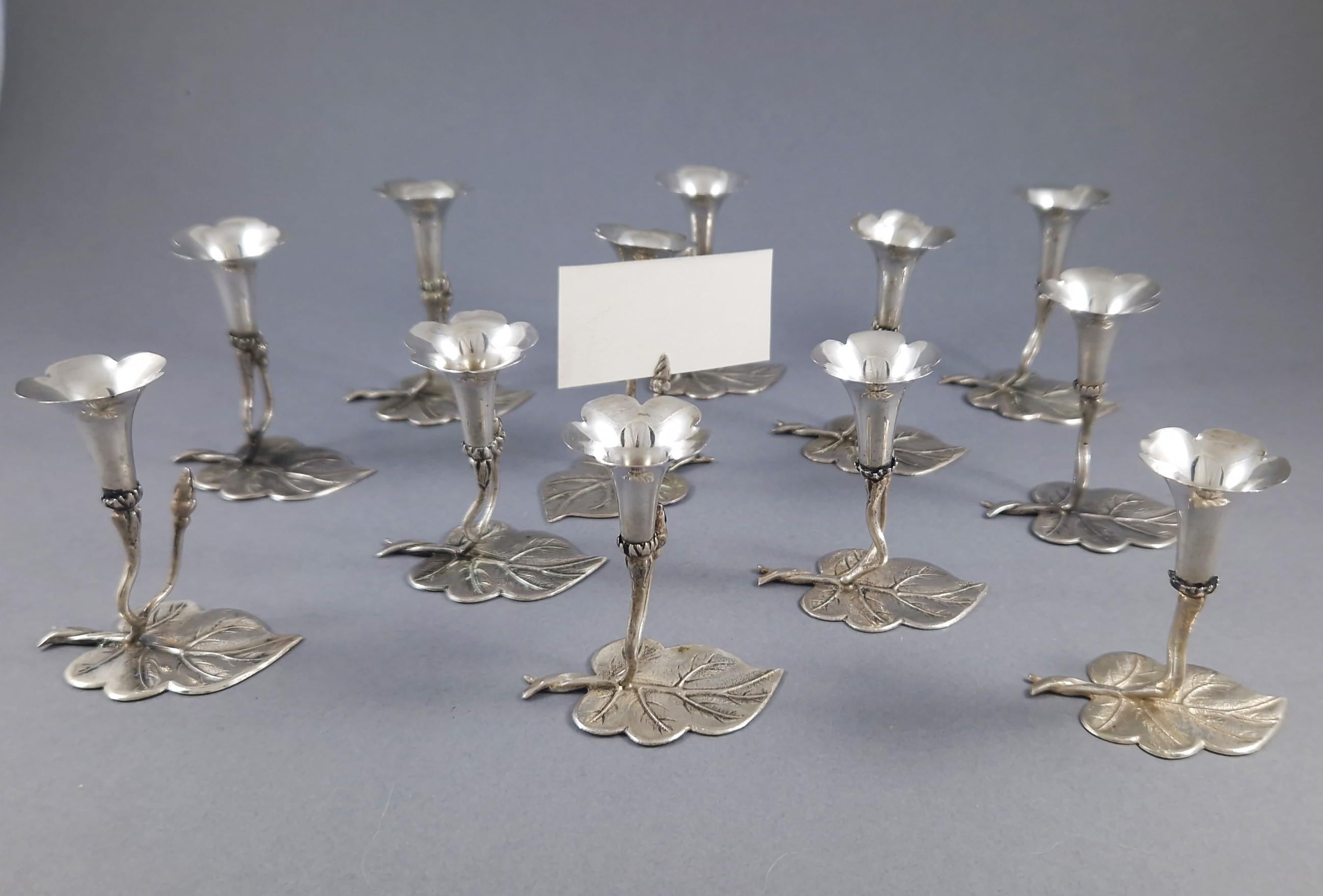 Zwölf silberne Tischkartenhalter, jeder in Form einer zarten Blume, die sich von einem blattförmigen Sockel erhebt. Die ausgestellte Blüte ist so gestaltet, dass sie entweder eine Tischkarte oder ein kleines Sträußchen aufnehmen kann.

800