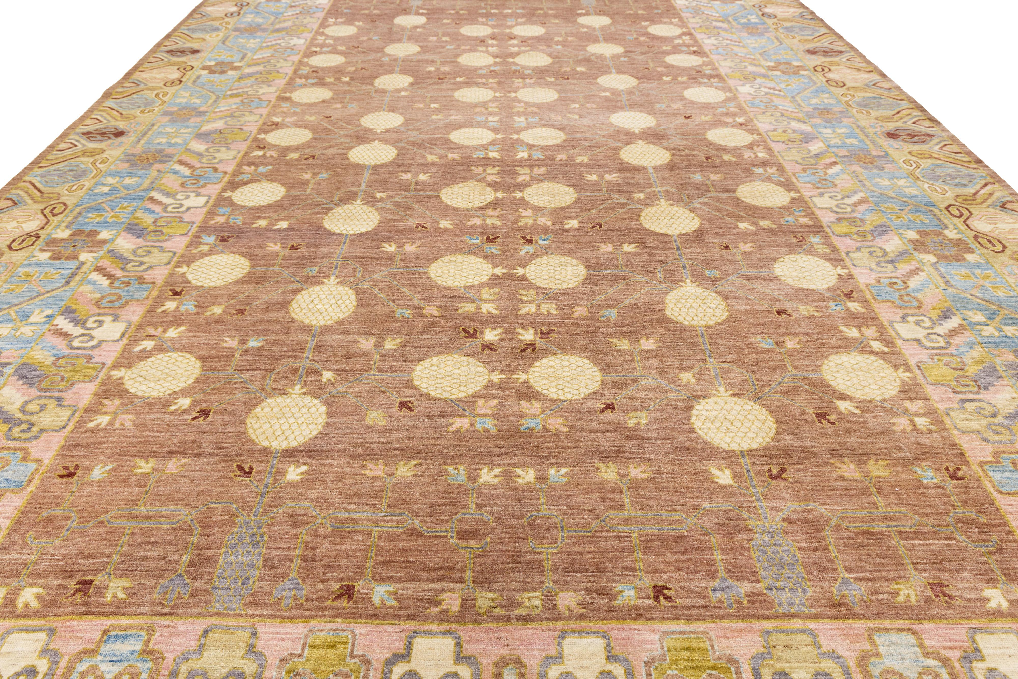 This Khotan-style hand-knotted wool rug features a warm brown field that is elegantly complemented by subtle accents of gold, pink, beige, and blue. Its design showcases a complex all-over interlock pattern, effectively merging traditional