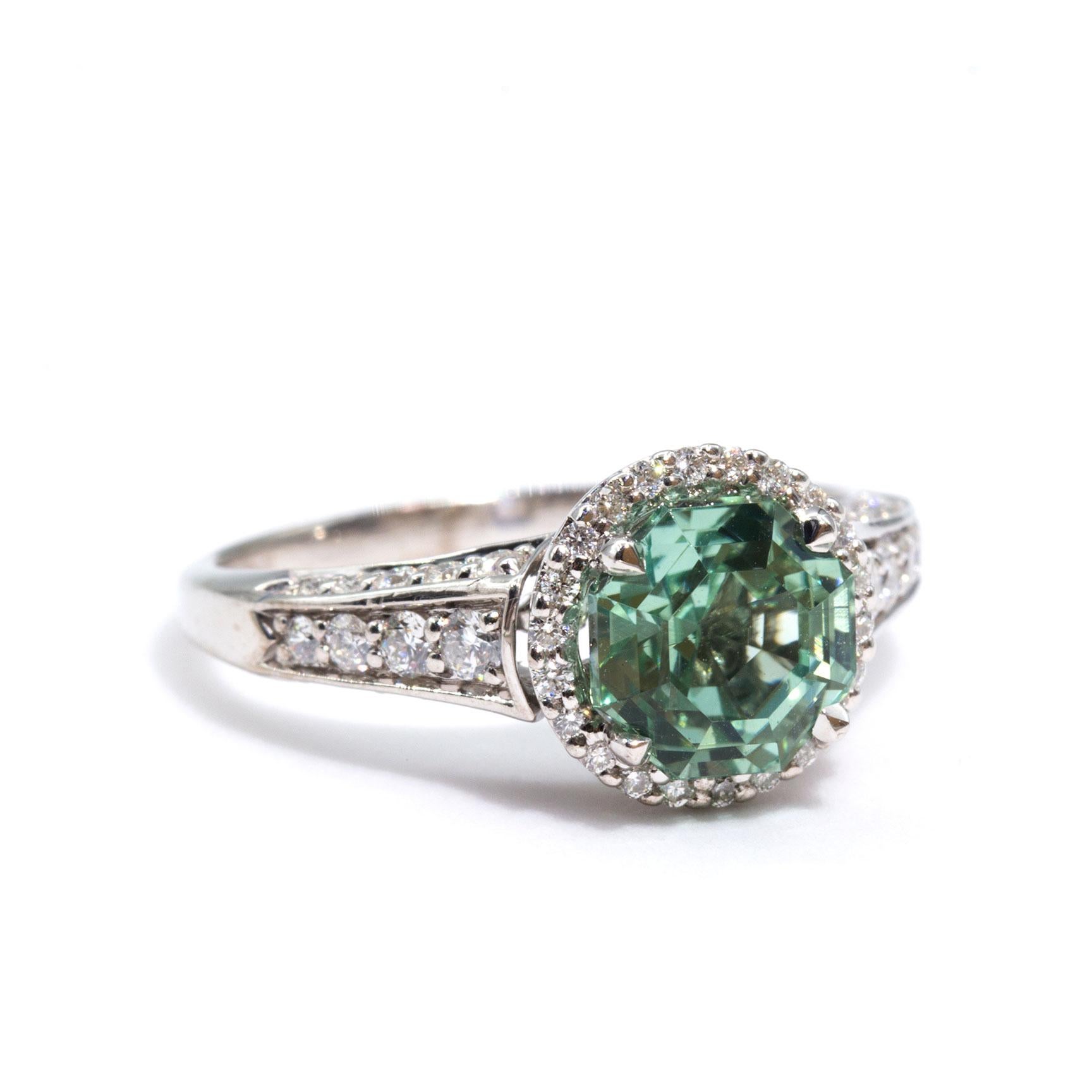 Crafted in 18 carat white gold is this unique vintage ring boasting a glorious 1.20 carat bright mint colour tourmaline and is encompassed by a halo of sparkling diamonds and is embellished with further round brilliant cut diamonds in the band. We