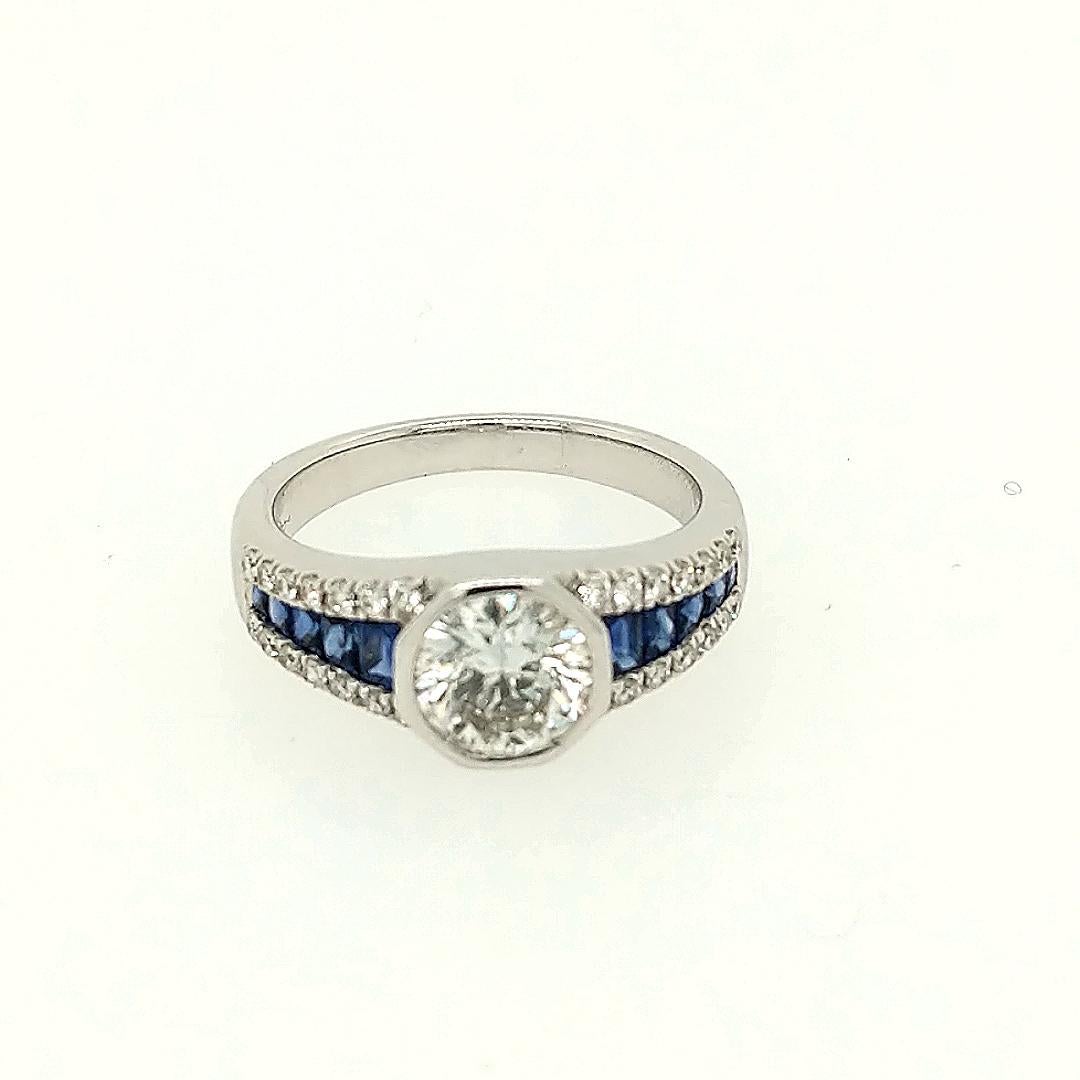 Crafted in 18k white gold featuring (1) brilliant round diamond weighing 1.20ct with a color of H and a clarity of I1. The I1 clarity is very good, looks eye clean. The diamond is bezel set and has (10) sapphire baguettes going down the side