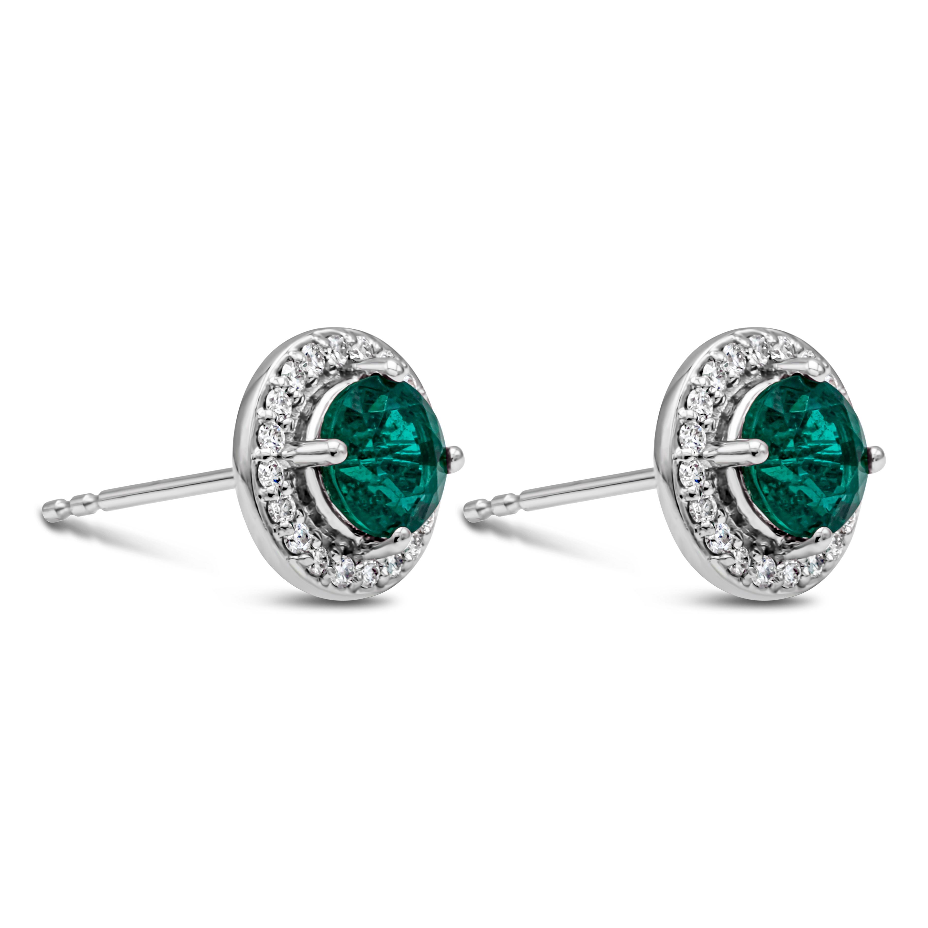 A simple and versatile pair of stud earrings showcasing 1.20 carats total round cut green emerald, set in a four prong basket setting. Surrounded by a single row of brilliant round diamonds weighing 0.21 carats total, F Color and VS in Clarity.