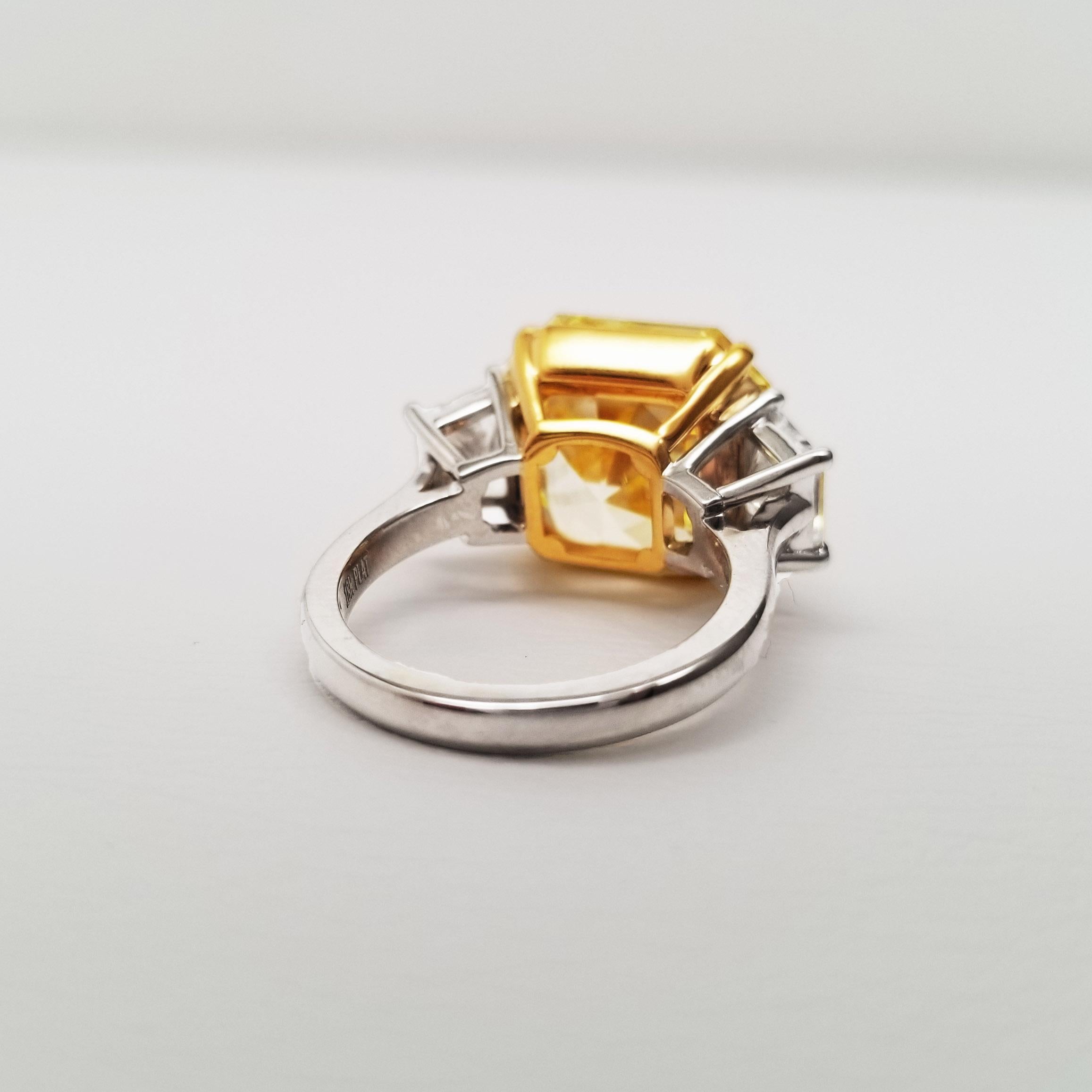 Three-Stone engagement ring from Scarselli featuring a 12.22 carat Fancy Intense Yellow center stone, Asscher-cut and GIA certified. The center stone has VVS1 clarity and is between two white, trapezoid cut side stones – 1.53 collective weight and