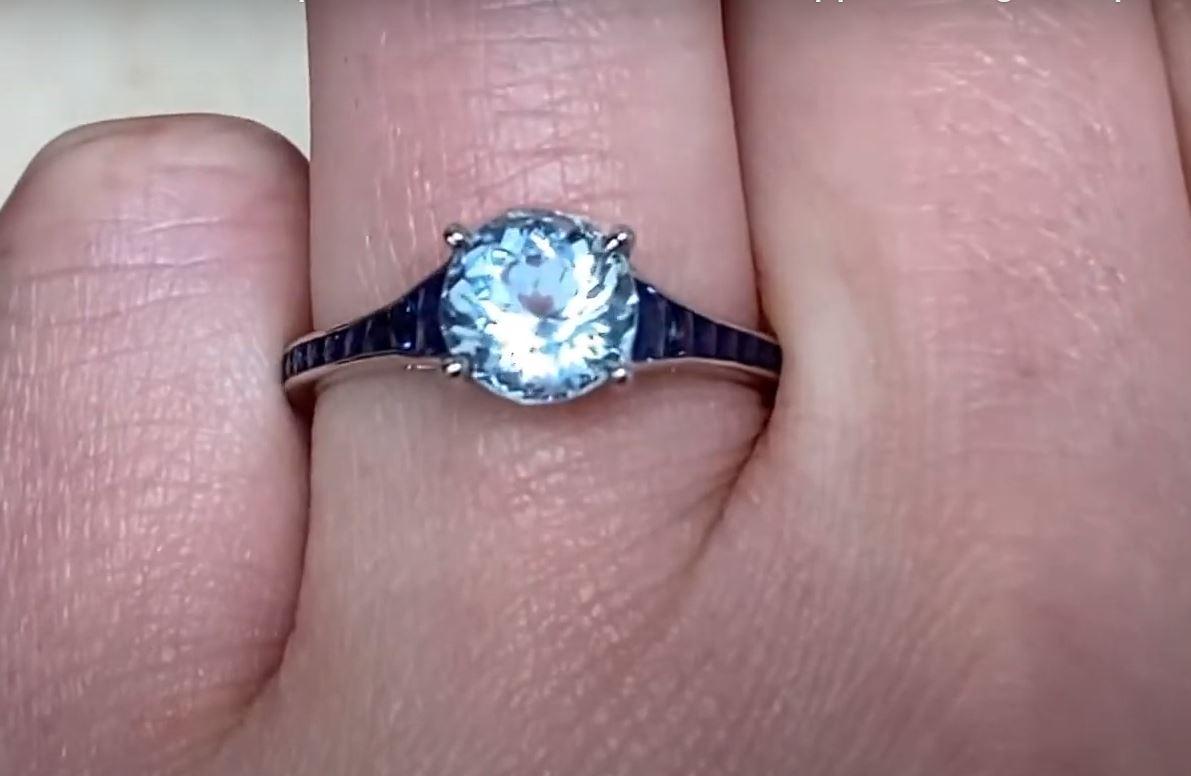 Dieser exquisite Ring zeigt einen leuchtenden Aquamarin im Rundschliff mit einem Gewicht von etwa 1,20 Karat. Sein bezaubernder Blauton wird durch eine Reihe von tiefblauen Saphiren, die sorgfältig entlang der Schultern gefasst sind, wunderbar