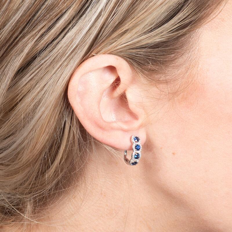 A beautiful huggie hoop earring featuring 1.25ct total weight in round blue sapphires surrounded by 0.26ct total weight in round cut diamonds set in 18K white gold. Lever back.