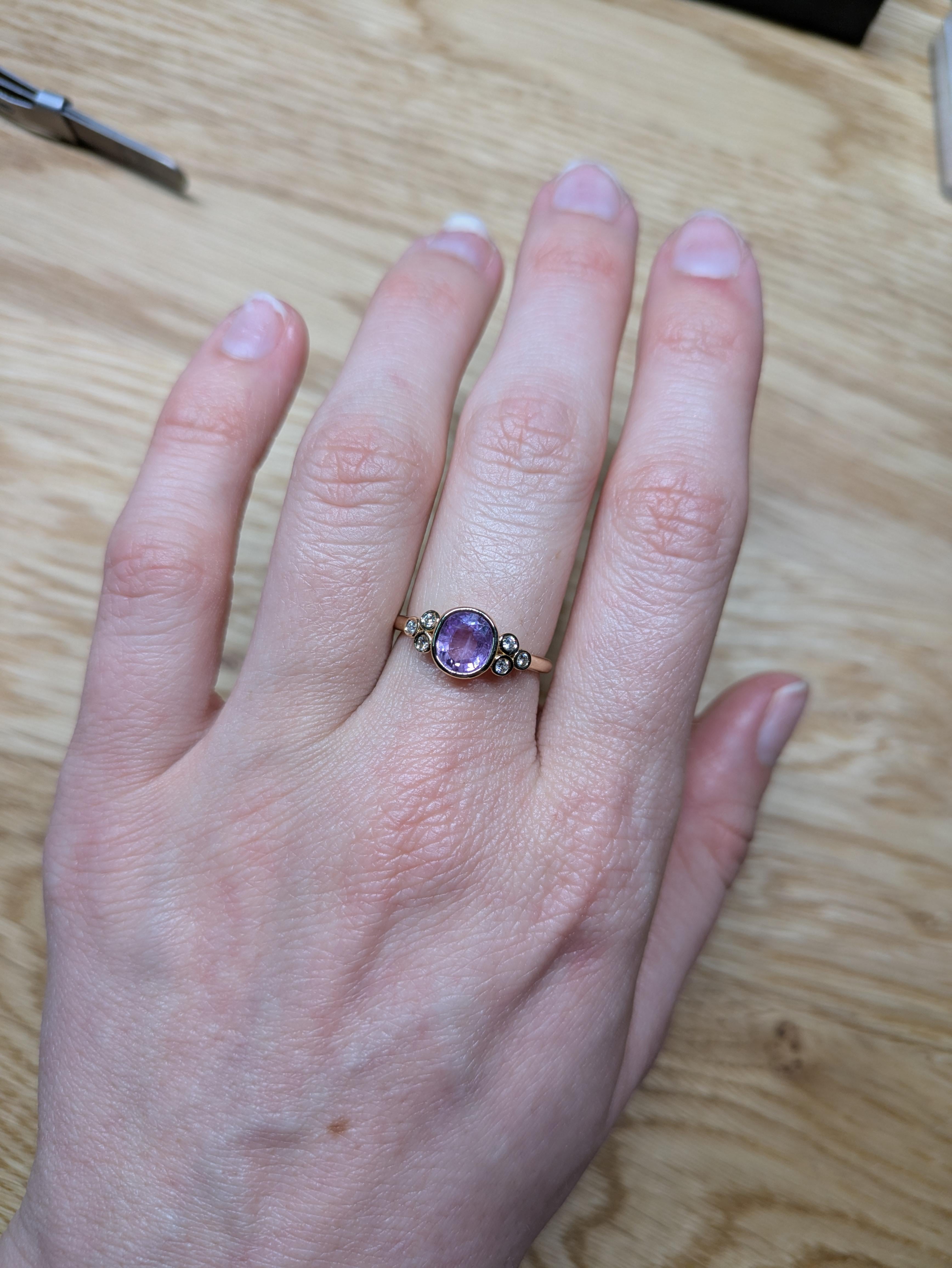 This ring features a stunning 1.26-carat oval-cut purple sapphire (7x6mm) set in 14K yellow gold. The sapphire is bezel-set and complemented by a total of 0.13 carats of diamonds, arranged in a cluster on either side of the center stone. The