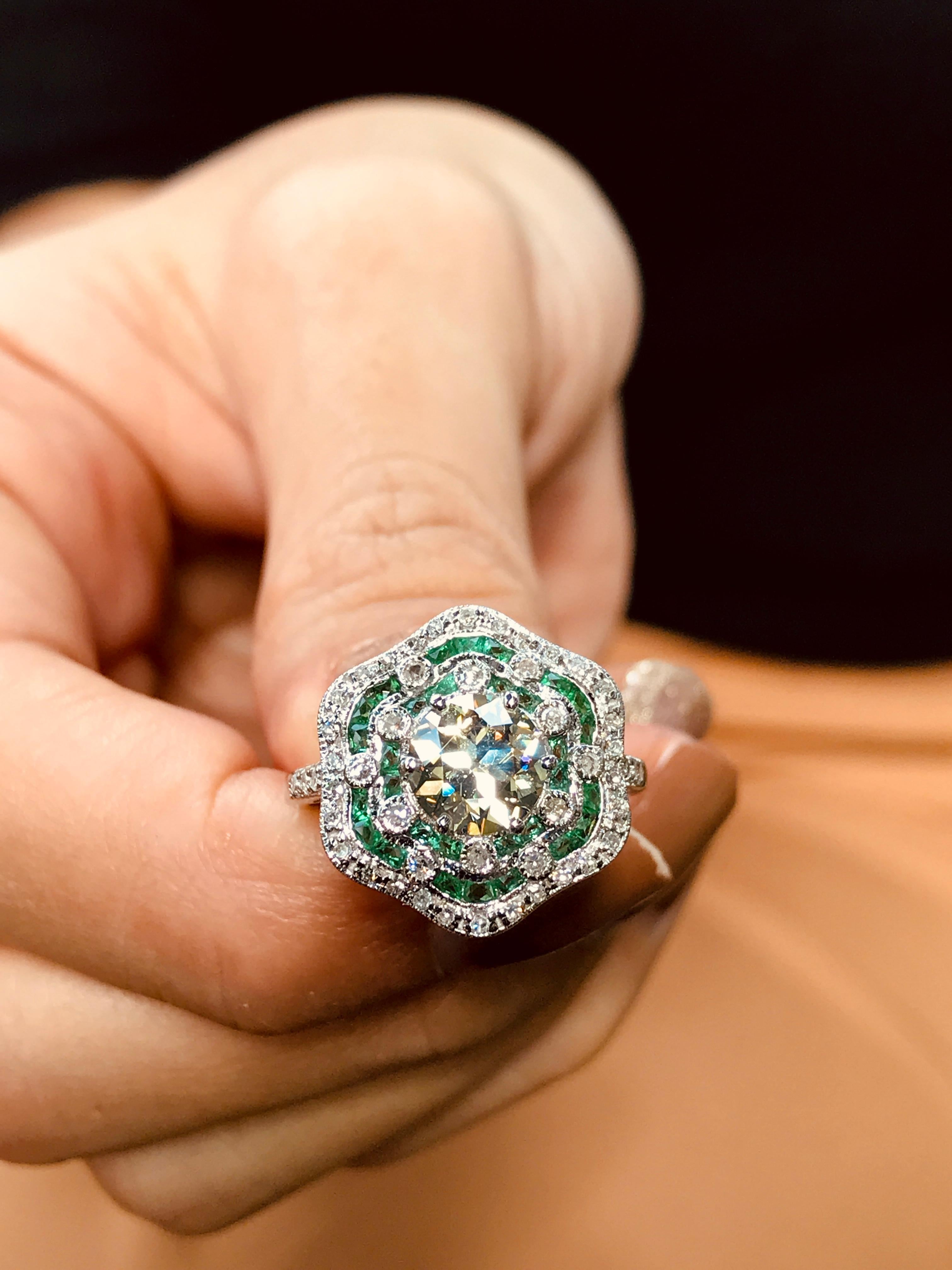 An elegant Art Deco inspired engagement ring is set with an antique old cut fancy yellow diamond and adorned with diamonds and millgrain. The diamond surrounded with vibrant green emeralds accented with round brilliant cut diamonds. Handcrafted in