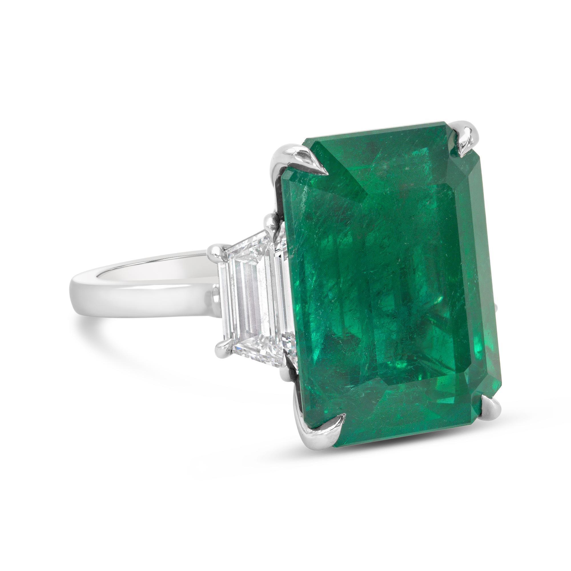 An elegant engagement ring, showcasing a C. Dunaigre certified 12.82 carats emerald cut green emerald, set on a classic four prong setting. Accented by two trapezoid cut diamonds weighing 1.02 carats total with D-E color and VS+ clarity. Elegantly