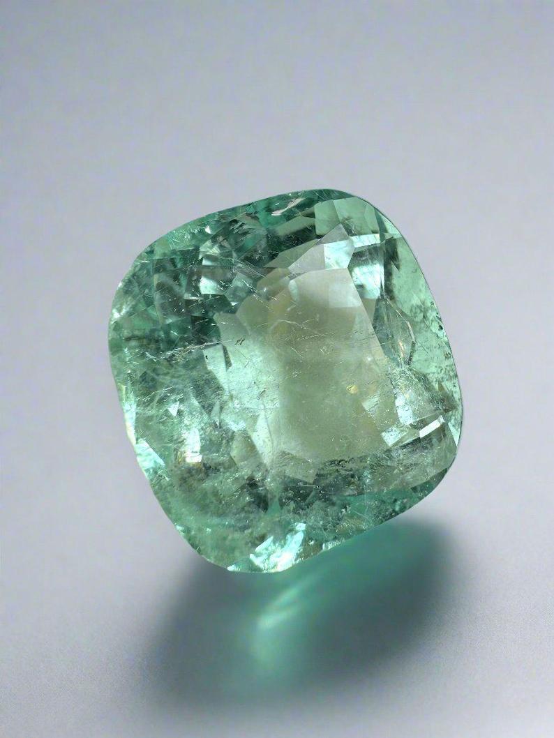 Stone: Emerald
Weight: 12.86-carats
Shape: Cushion Cut
Clarity: Transparent
Luster: Very Good
Color: Pastel Bluish Green
Measurements: 14.51mm x 14.26mm x 9.80mm
Geographic Origin: Colombia
Treatment: Natural, Oil

Comments: Due to their growth