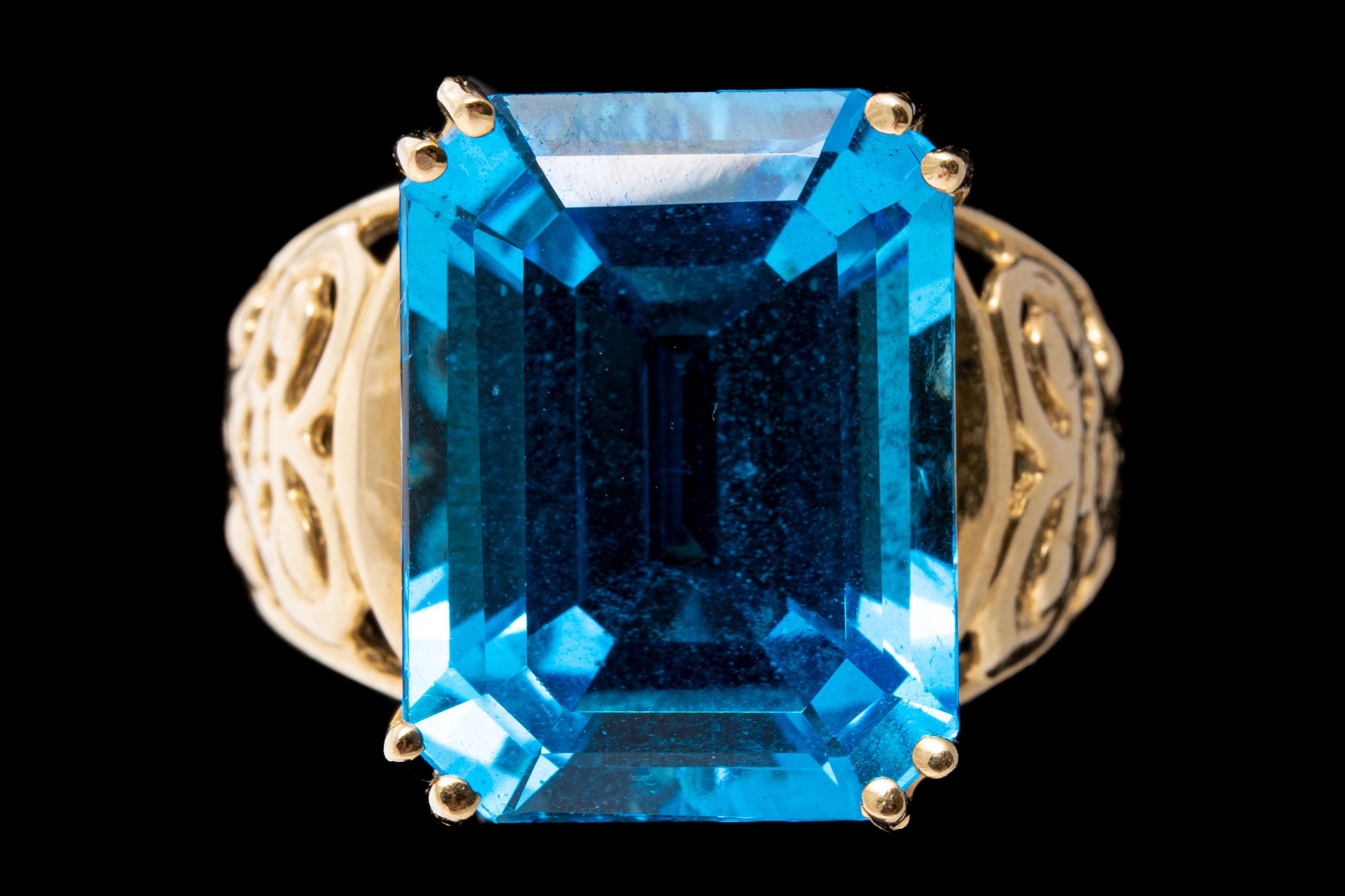 12k yellow gold ring. This striking ring has a wide, emerald cut faceted, deep blue color blue topaz, approximately 13.21 CTS, split prong set and decorated with wide, pierced shoulders.
Marks: None, tests 12k
Dimensions: 7/16