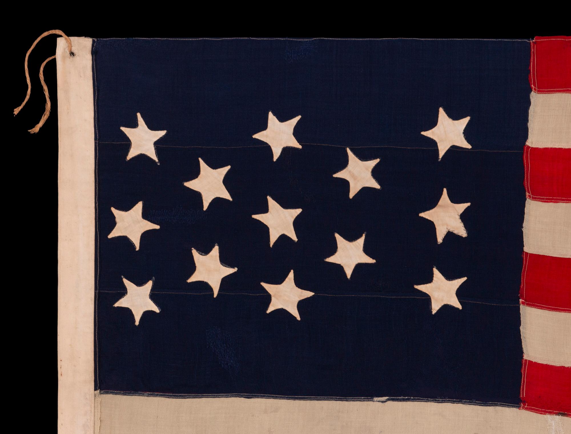 13 STAR ANTIQUE AMERICAN FLAG WITH HAND-SEWN STARS IN AN EXTREMELY NARROW PRESENTATION OF A 3-2-3-2-3 ARRANGEMENT ON A CANTON THAT DOESN’T FOLLOW SUIT, LEAVING WIDE EXPANSES OF BLUE WOOL BUNTING ABOVE AND BELOW; MADE IN THE ERA OF THE 1876