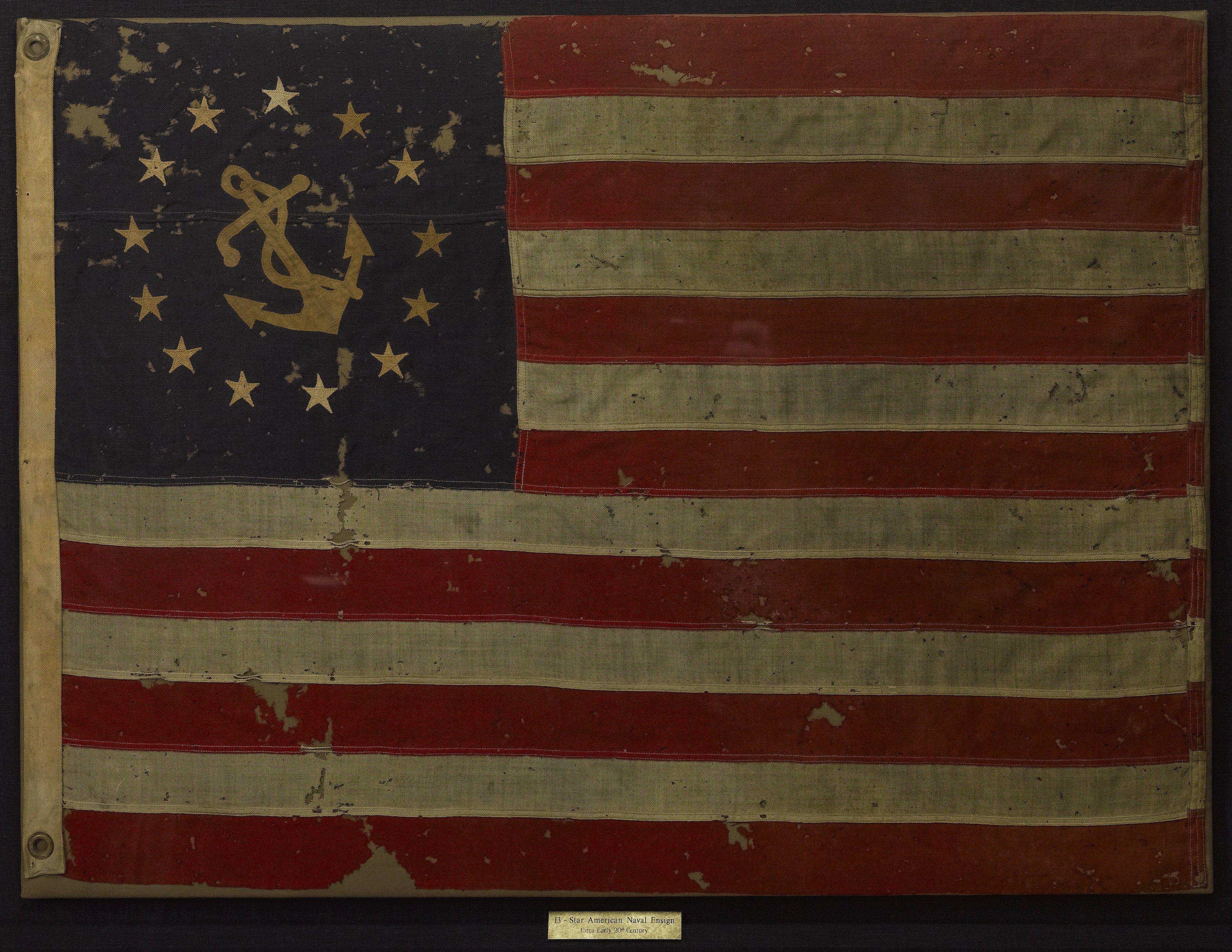 This is a 13-star American Naval Ensign from 1877. The flag has a large fouled anchor at the center of the canton. The anchor is encircled by 13 white stars. The circular arrangement of stars suggests unity and perpetuity, and it was a favorite
