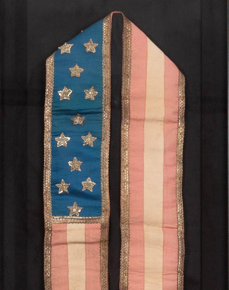 Presented is an original patriotic sash from the late 19th century, featuring 13 stars against a bright blue field. This sash features appliqued silver stars on a blue canton, red and white stripes, a round banner button, and marking on interior