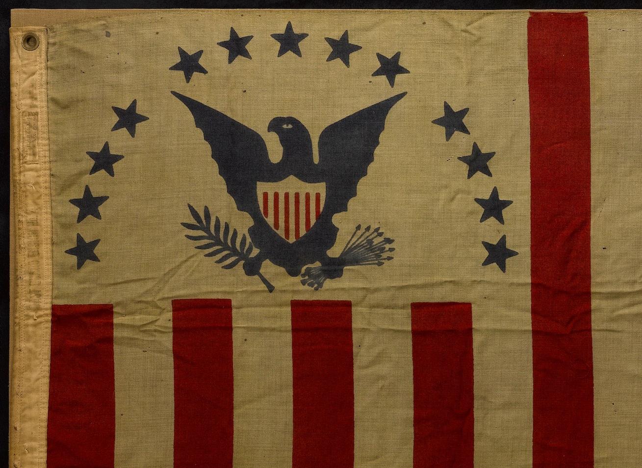 This is a United States Coast Guard Flag, circa 1895-1930. The flag was machine printed on cotton. Ensign features an eagle on the canton, with spread wings and an olive branch in one talon with a bunch of arrows in the other. Above the eagle on the