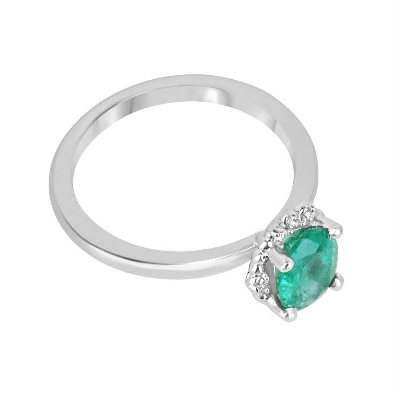 Presenting our Emerald and Diamond Ring, a symbol of elegance and grace. At its heart lies a captivating 1.20-carat oval cut emerald, boasting a lush medium dark green tone and semi-transparent clarity, measuring a rounded 8 x 6 mm. Encircled by a