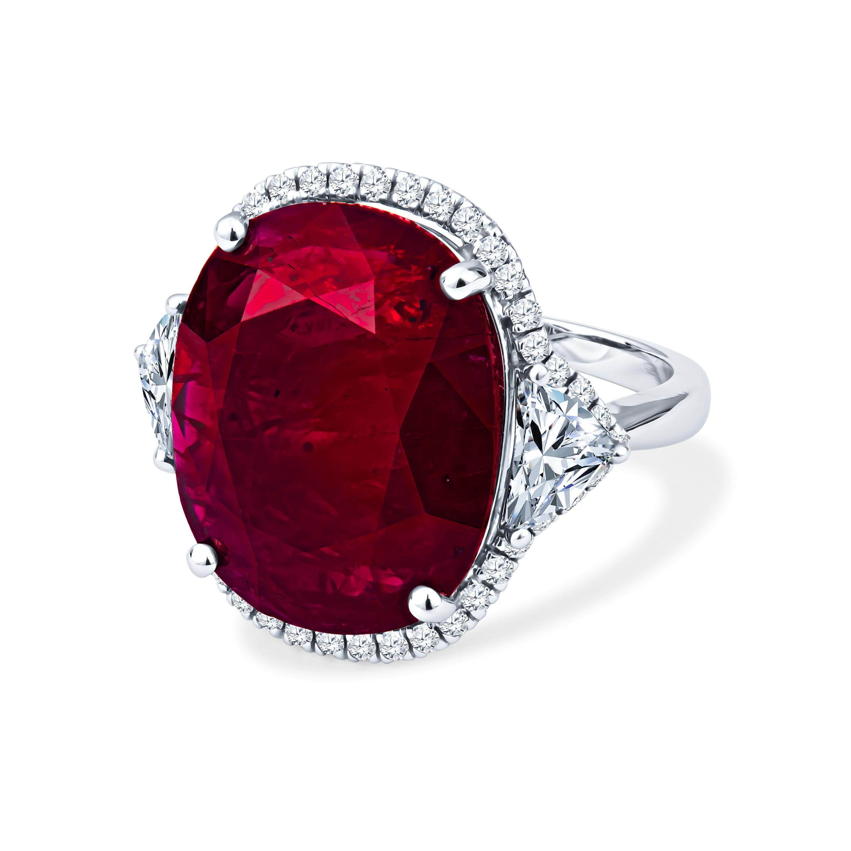 This ring showcases a 13.12ct oval cut natural ruby (standard heat treatment only), surrounded by 0.44ct round diamonds forming a diamond halo and 2 0.82ctw trillion cut diamonds in the sides. The ring is set in 18kt white gold and is a size 6.5,