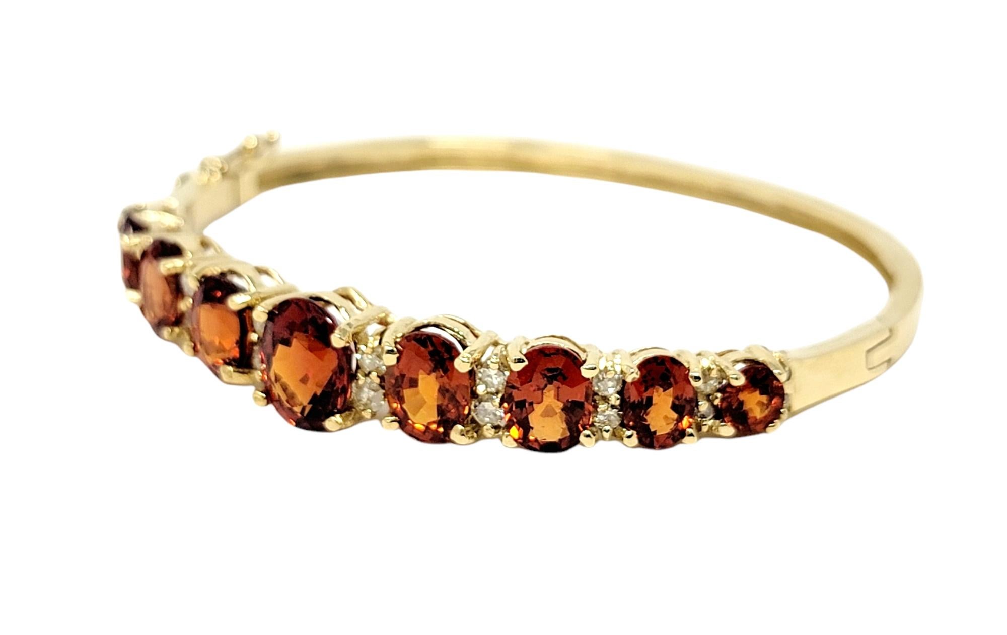 Sleek and elegant yellow gold bangle bracelet with colorful sparkling spessartine garnet and diamond detailing. This chic hinged bangle features graduated bright reddish-orange garnets set in a single row accented by dazzling diamonds in between