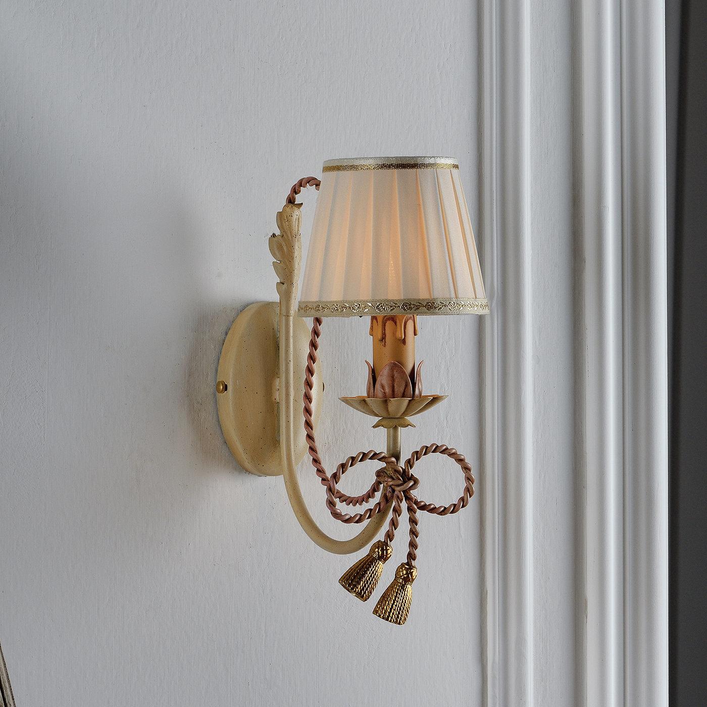 Decorado a mano y con detalles en pan de oro, este pequeño aplique en forma de lámpara está sujeto por una pieza de pared de color marfil y tiene un delicado cordón metálico con borlas atadas en un lazo. Una sola bombilla está cubierta por una