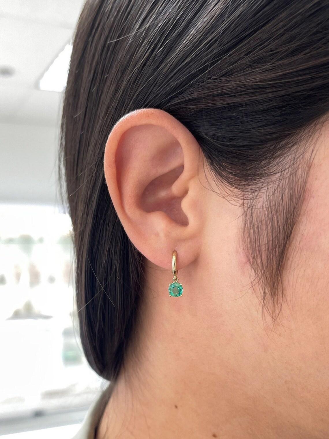 These stunning emerald dangle earrings boast a combined weight of 1.35 carats of round-cut emeralds, elegantly showcased in a four-prong basket setting. Suspended from a 10mm knife edge huggie style crafted in luxurious 14k gold, they offer a