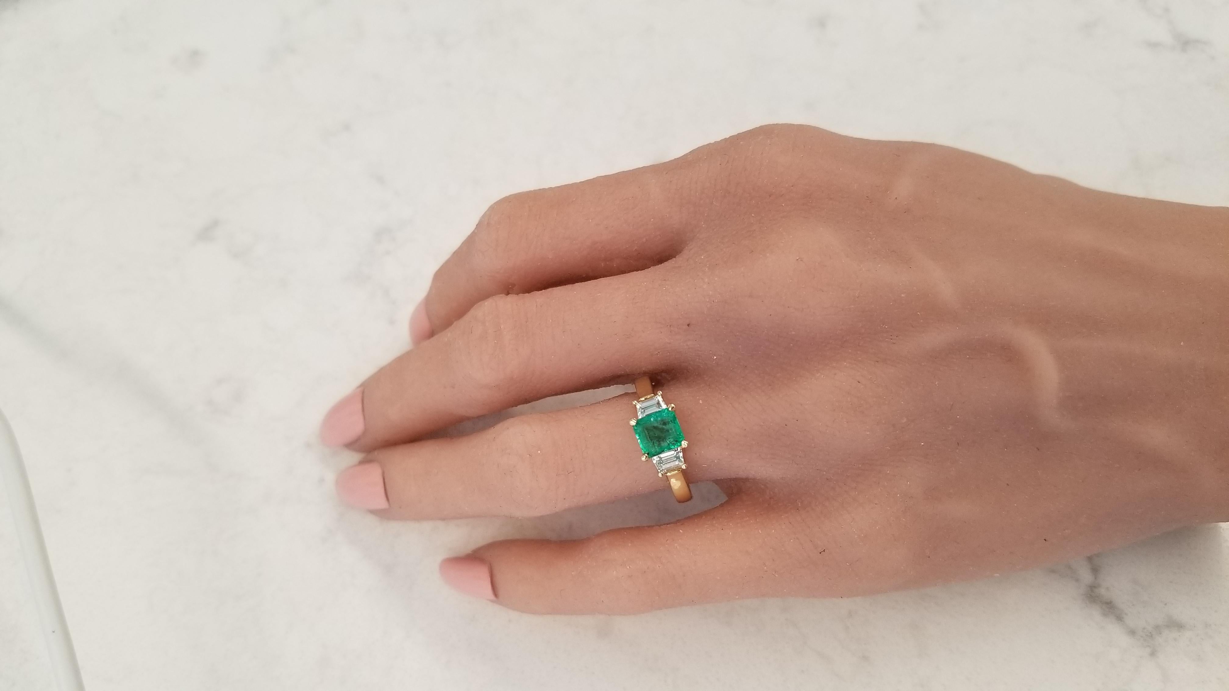This three stone ring showcases a 1.37 carat square emerald cut green emerald, prong set, that measures 6.2x5.8MM. The gem source is Zambia; its color is evenly distributed throughout the gem; its luster and transparency are excellent. This emerald