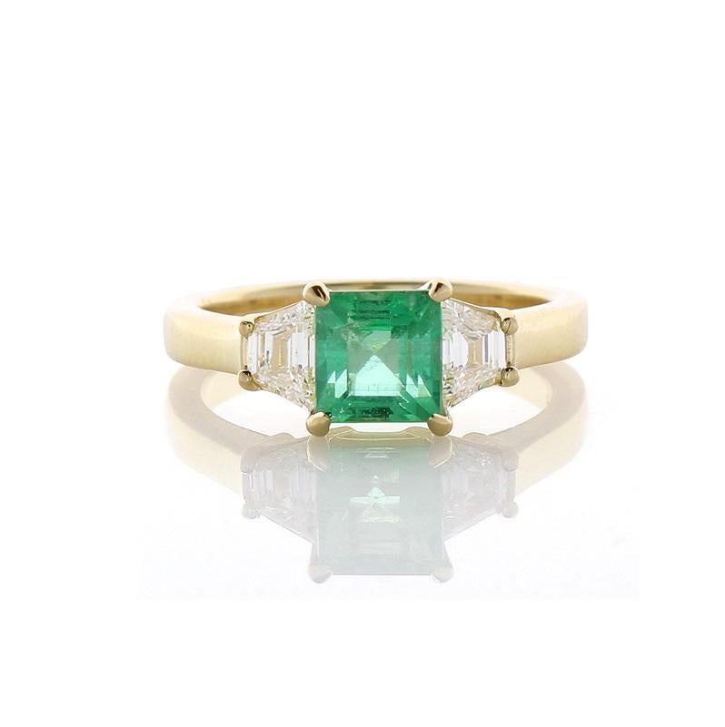 This three stone ring showcases a 1.38 carat square emerald cut green emerald, prong set, that measures 5.8x6.1MM. The gem source is Zambia; its color is evenly distributed throughout the gem; its luster and transparency are excellent. This emerald