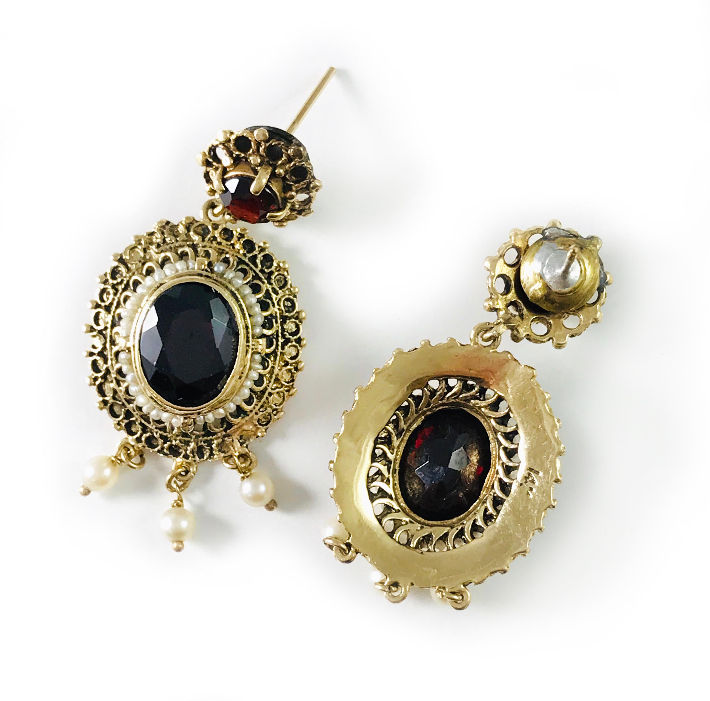 14 Karat Antique Garnet Seed Pearl Earrings. An ornate design with an oval and round garnet and three petite pearls that dangle on the bottom of each earring. A single row of seed pearls surround the oval bezel-set Garnet. The romantic style make