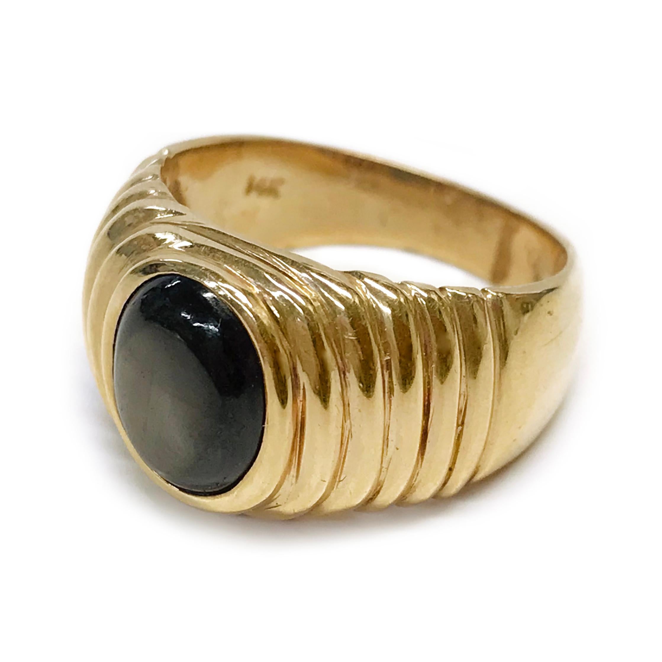 14 Karat Black Star Sapphire Ring. The ring features a bezel-set oval cabochon black star sapphire. The sapphire is 3.34ct and the ring size is 8. Approximately half of the ring band is a smooth shiny finish with smooth raised ridges radiating from