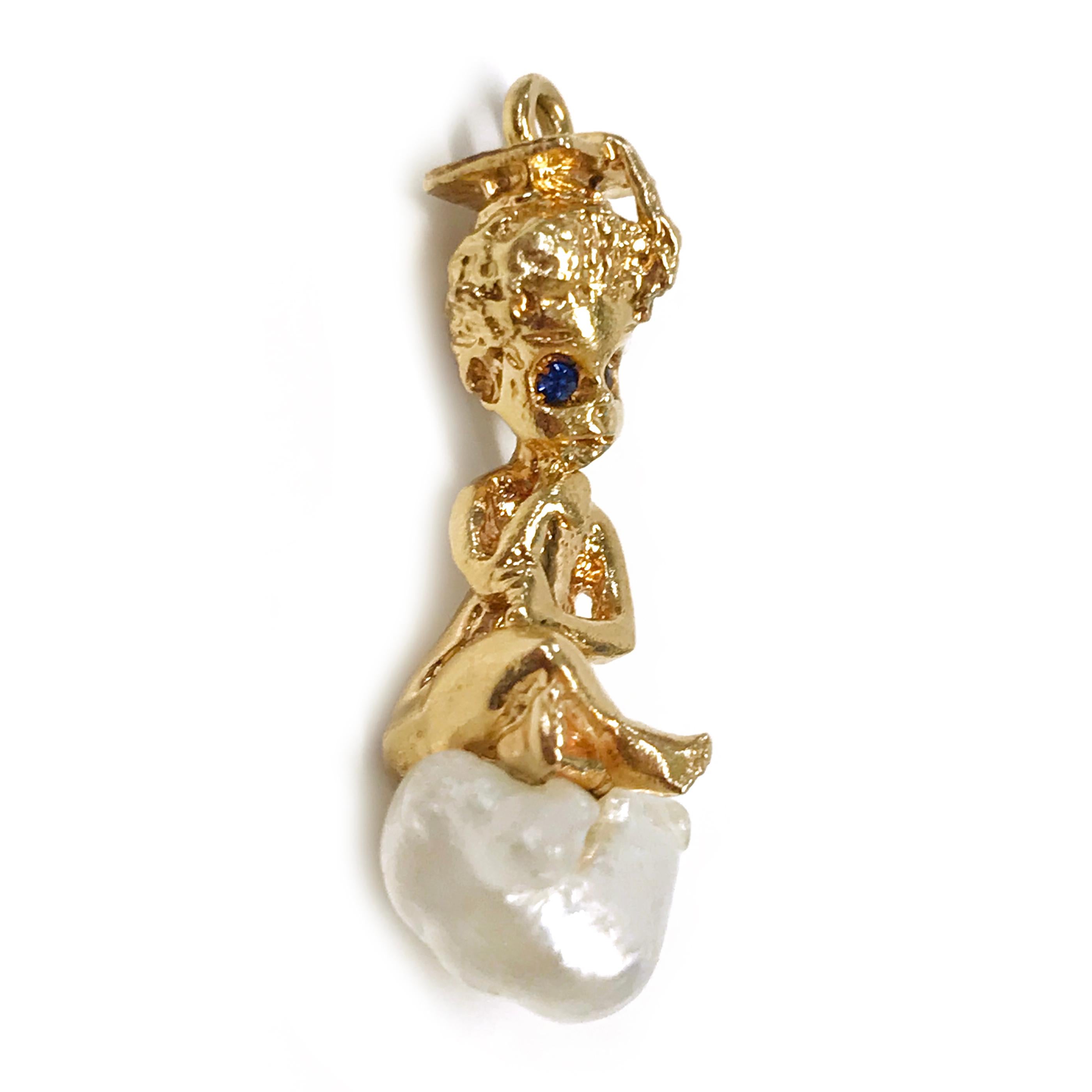 14 Karat Graduation Boy Blue Sapphire Pearl Pendant. This cute little boy is wearing a graduation cap, has blue sapphire eyes, and is sitting on a cloud-shaped Baroque pearl. The overall texture is smooth with detail in the curly hair. The pendant