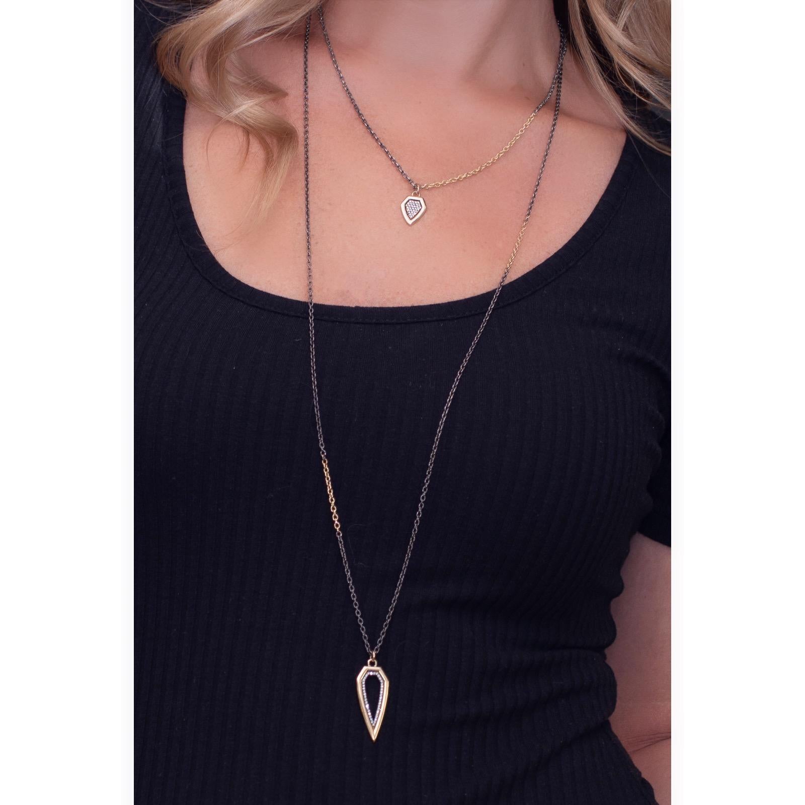 Un ciondolo moderno Ispirato alle forme degli scudi vittoriani, questo elegante ciondolo presenta una fila di diamanti lungo il bordo interno.  È appeso a una lunga catena in argento annerito e oro 14 carati.  
Materiali: Oro 14 carati, diamanti da