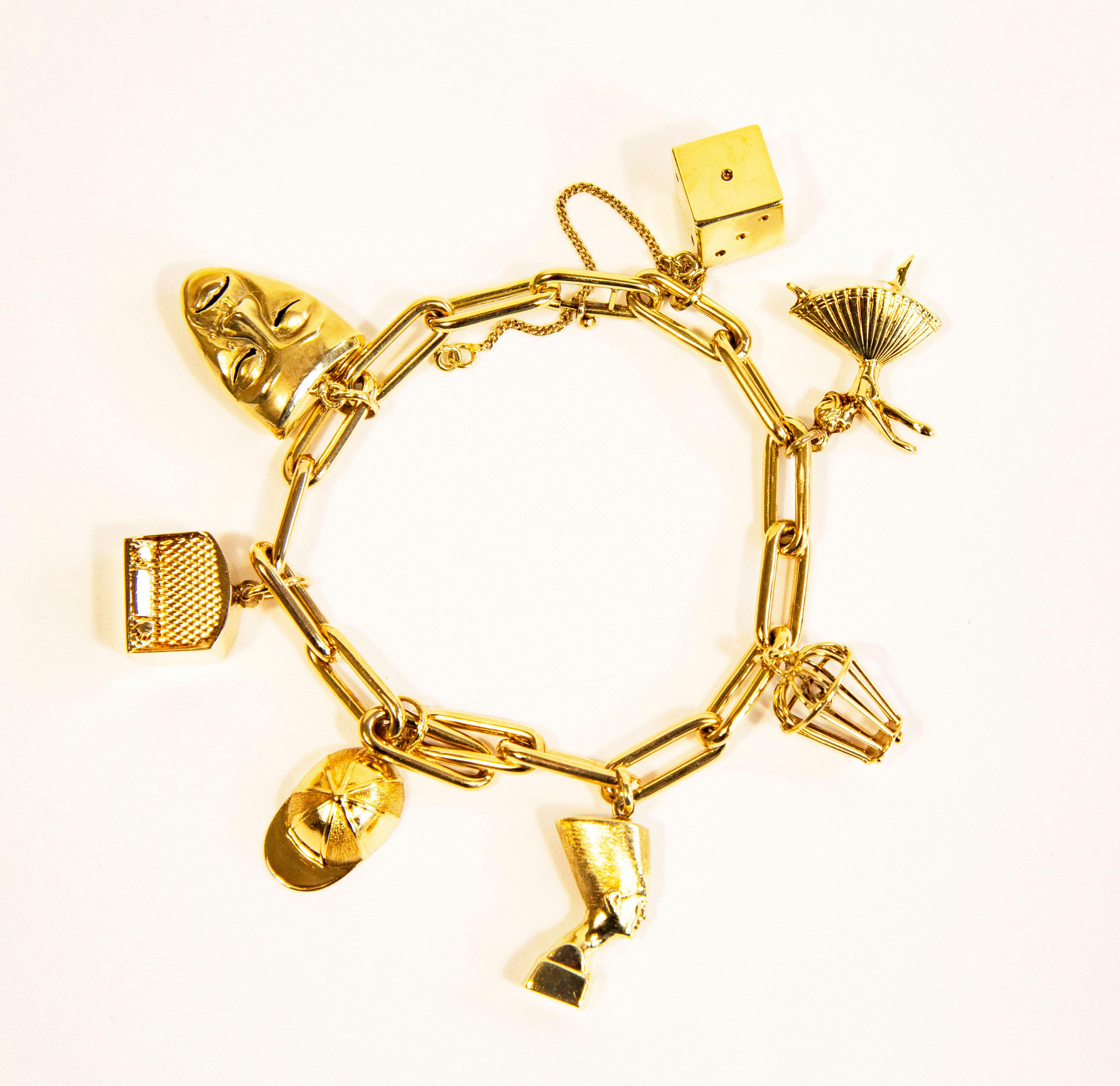 A vintage 14 karat yellow gold chain bracelet with 7 charms, including a dice, a ballerina, a closed basket/cage with a heart inside, Nefertiti, a cap, a radio, and an actor's mask. The bracelet features a safety chain attached to the distal links.