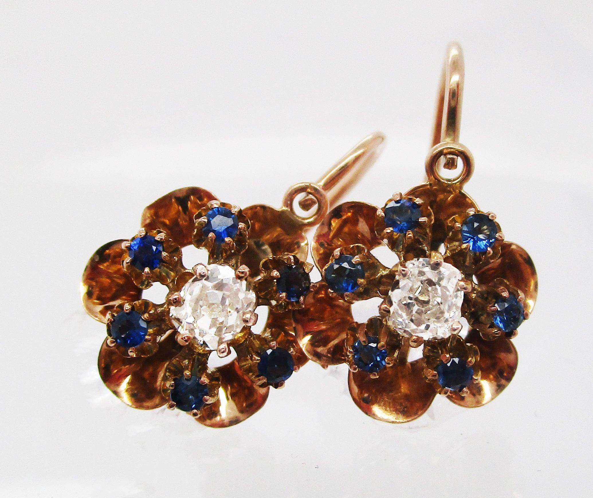 This is a stunning pair of Victorian drop earrings in 14k rose gold boasting gorgeous mine cut diamond centers framed by brilliant blue sapphires set into a beautiful rose gold flower setting! The lovely design of the earrings features a blue