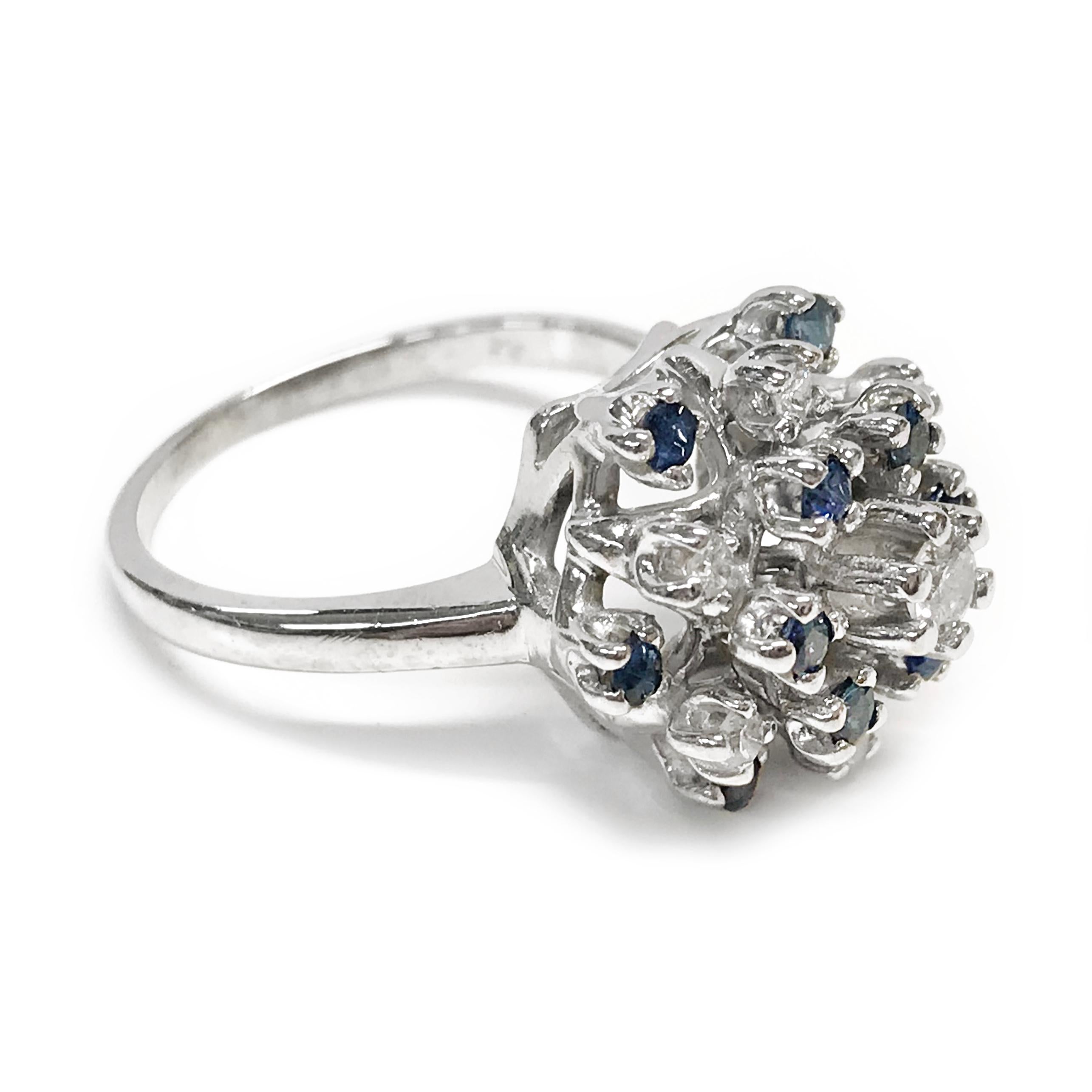 14 Karat White Gold Diamond Blue Sapphire Ring. The cluster ring features round diamonds and blue sapphires set in tiers of multiple heights. There is a total of seven diamonds, one 3.4mm at the center and six 0.018mm in a star shape. Twelve round
