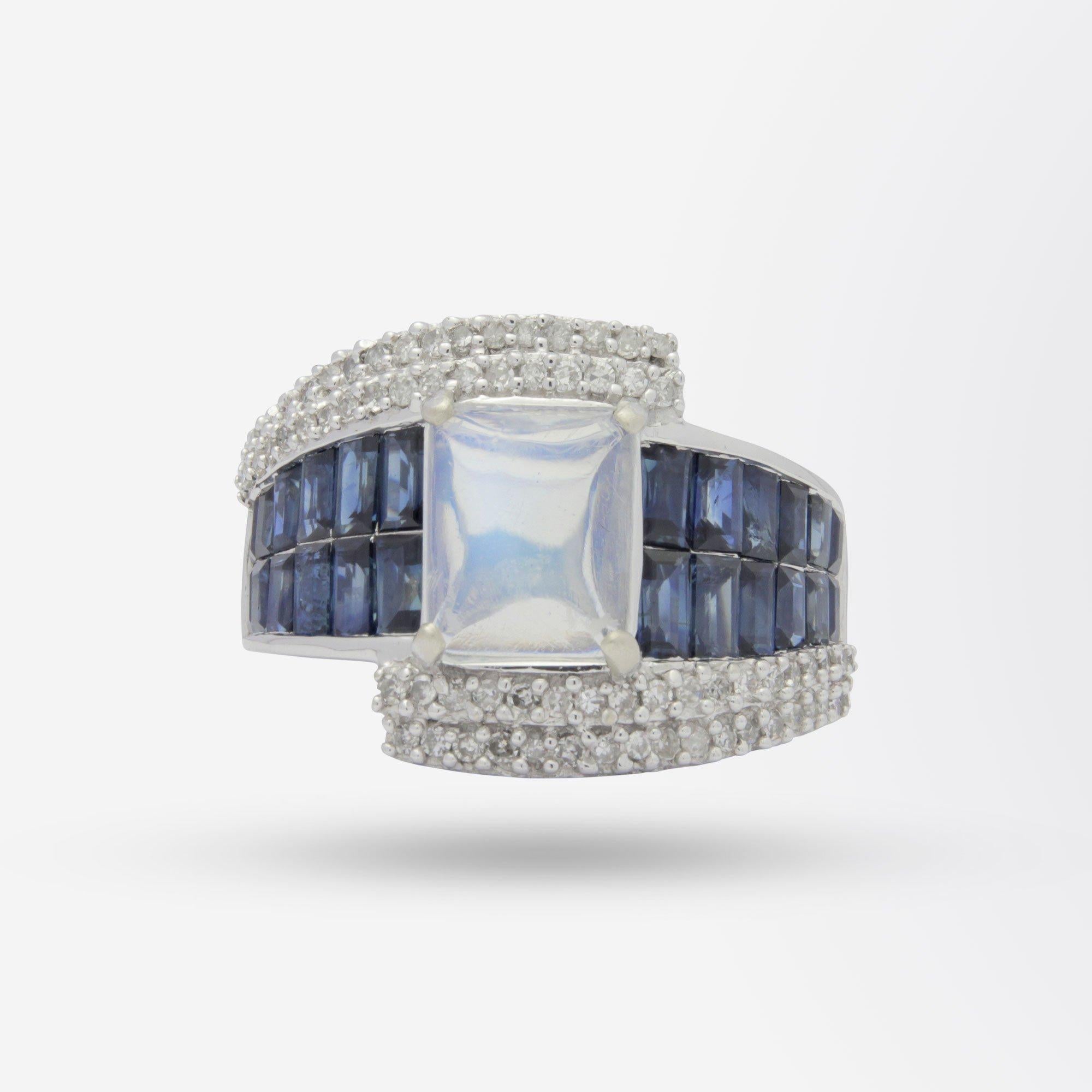 A fine 14 karat white gold ring set with a central moonstone and flanked with sapphires and diamonds. The cocktail ring features 93 stones in total and is asymmetric in design centring on a rectangular cabochon cut moonstone. The shoulders of the
