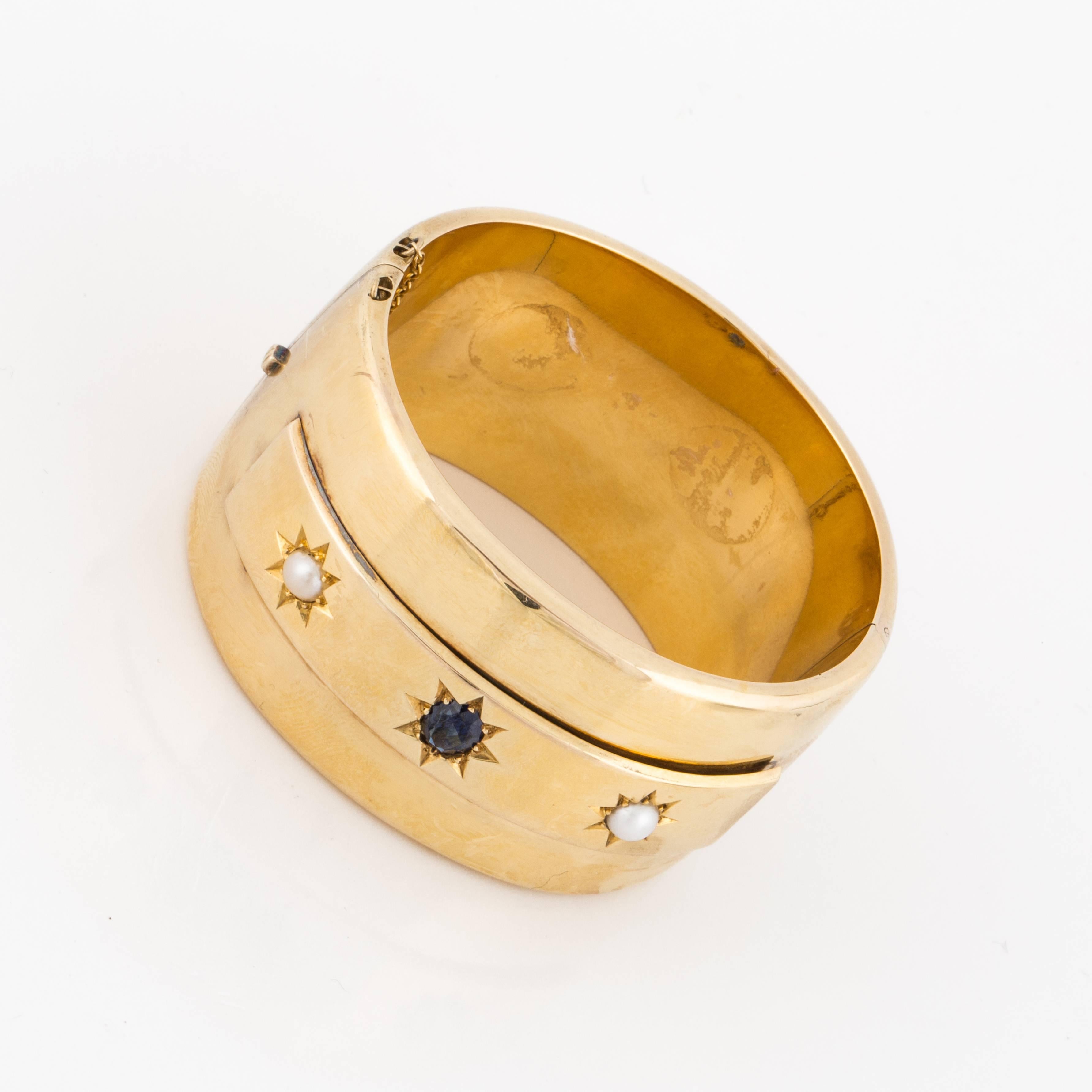 14K yellow gold wide bangle bracelet.  It has a smooth shiny polish and features an oval sapphire in the center with a pearl set on each side in a starburst design.  Measures 1-3/8