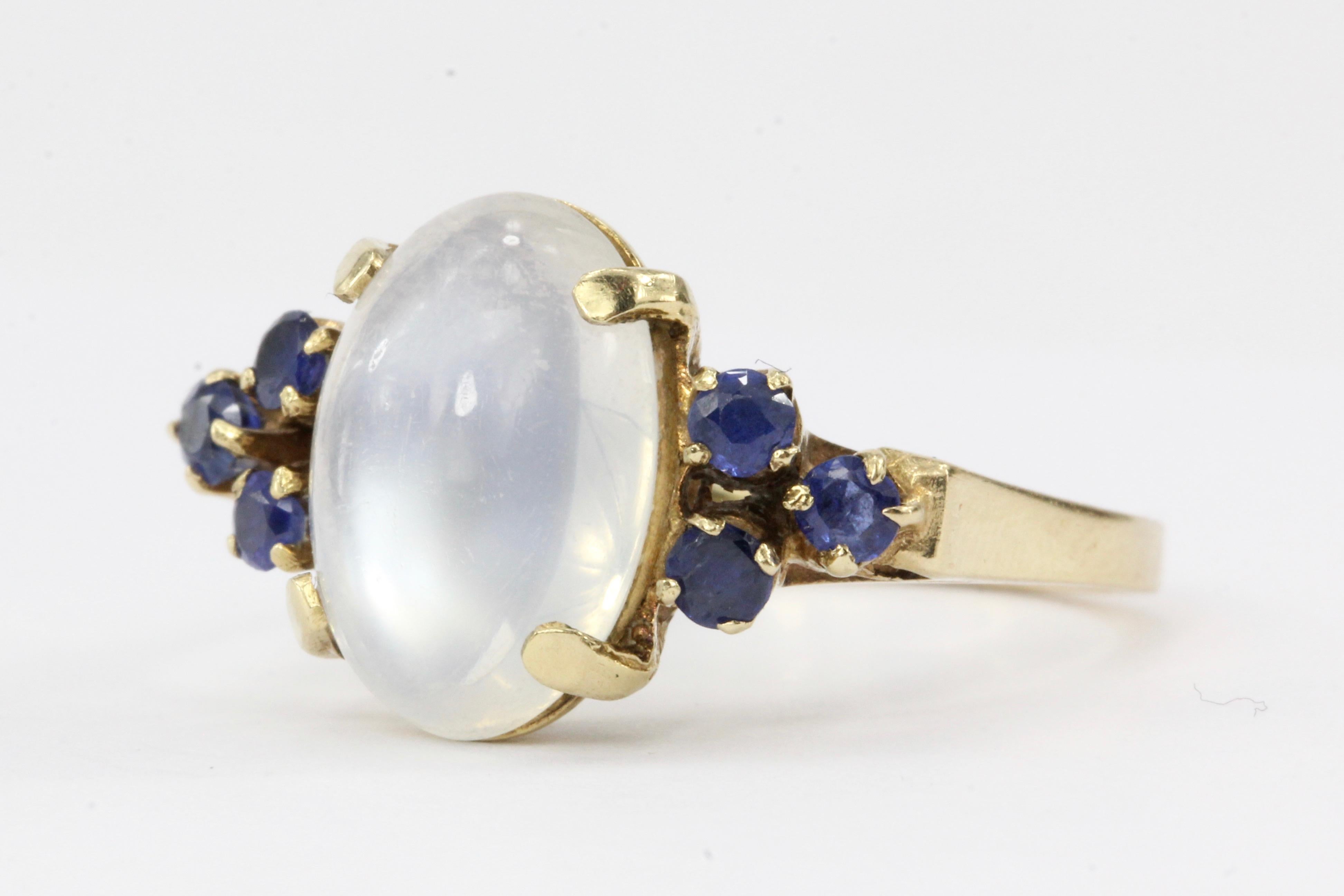 Era: Vintage

Hallmarks: 14K, WI

Composition: 14K Yellow Gold

Primary Stone: Moonstone

Stone Carat: Approximately 4 carats

Shape: Oval Cabochon

Accent Stone: Blue Sapphires

Total Carat Weight: Approximately .24 carats

Ring Width: