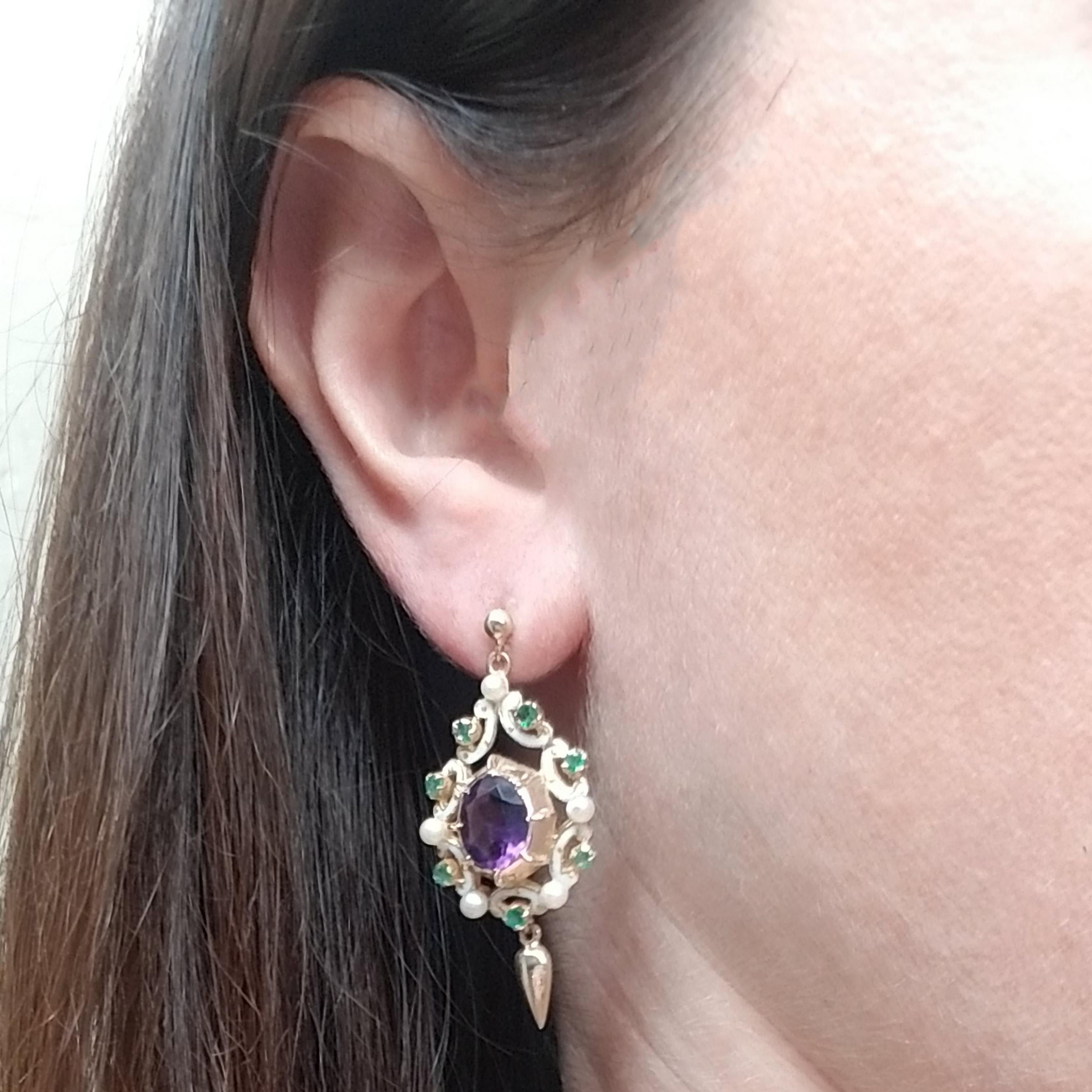 14 Karat Yellow Gold Dangle Earrings Featuring 2 Prong Set Oval Amethysts Totaling Approximately 4 Carats. The Center Stones are Complemented by 14 Prong Set Round Emeralds Totaling Approximately 0.42 Carats, 10 Cultured Pearls, & White Enamel