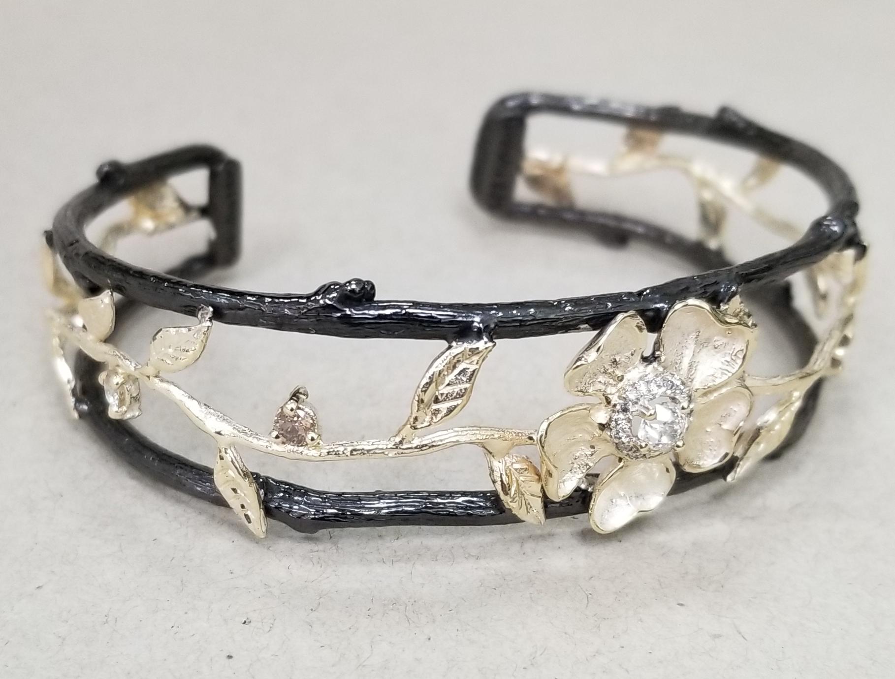14k yellow gold Gresha signature bark, flower and leaf diamond cuff, containing 25 round diamond; whites and brown weighing .85pts.  The  diamonds are paved around a .20pt. diamond.  The silver has been black rhodium for antiquing. 
*this design is